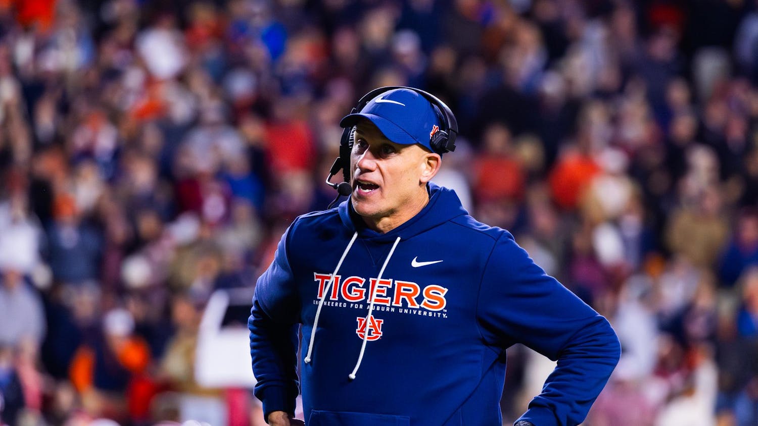 A man in a blue sweatshirt with "TIGERS" written on it stands on the sidelines of a football game, holding papers and wearing a headset.