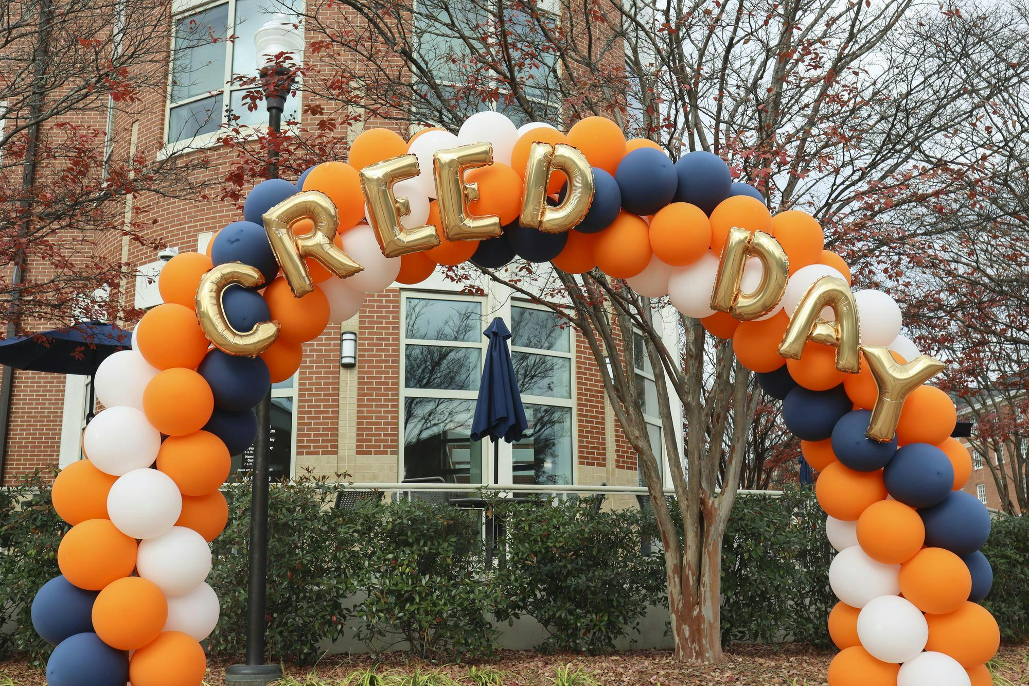 A colorful balloon arch with gold letters spelling "CREED DAY" stands near a brick building and leafless trees.