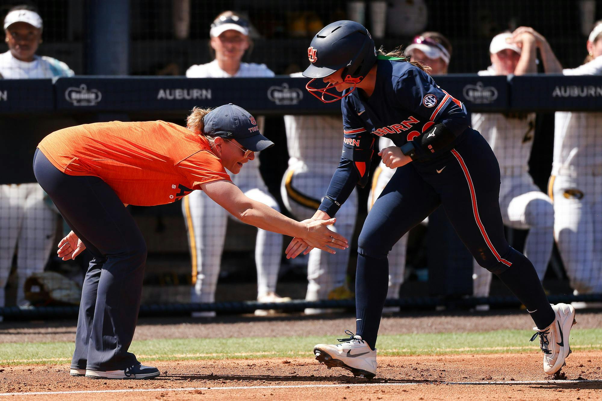 A coach in an orange shirt and cap extends her hand for a high five with a player in a blue uniform.
