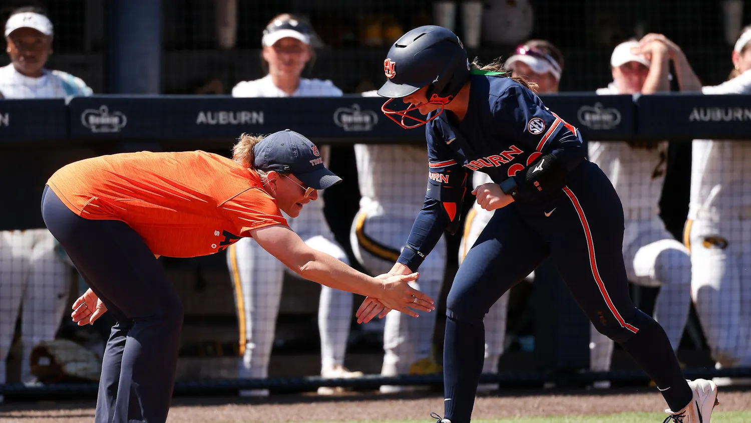 A coach in an orange shirt and cap extends her hand for a high five with a player in a blue uniform.