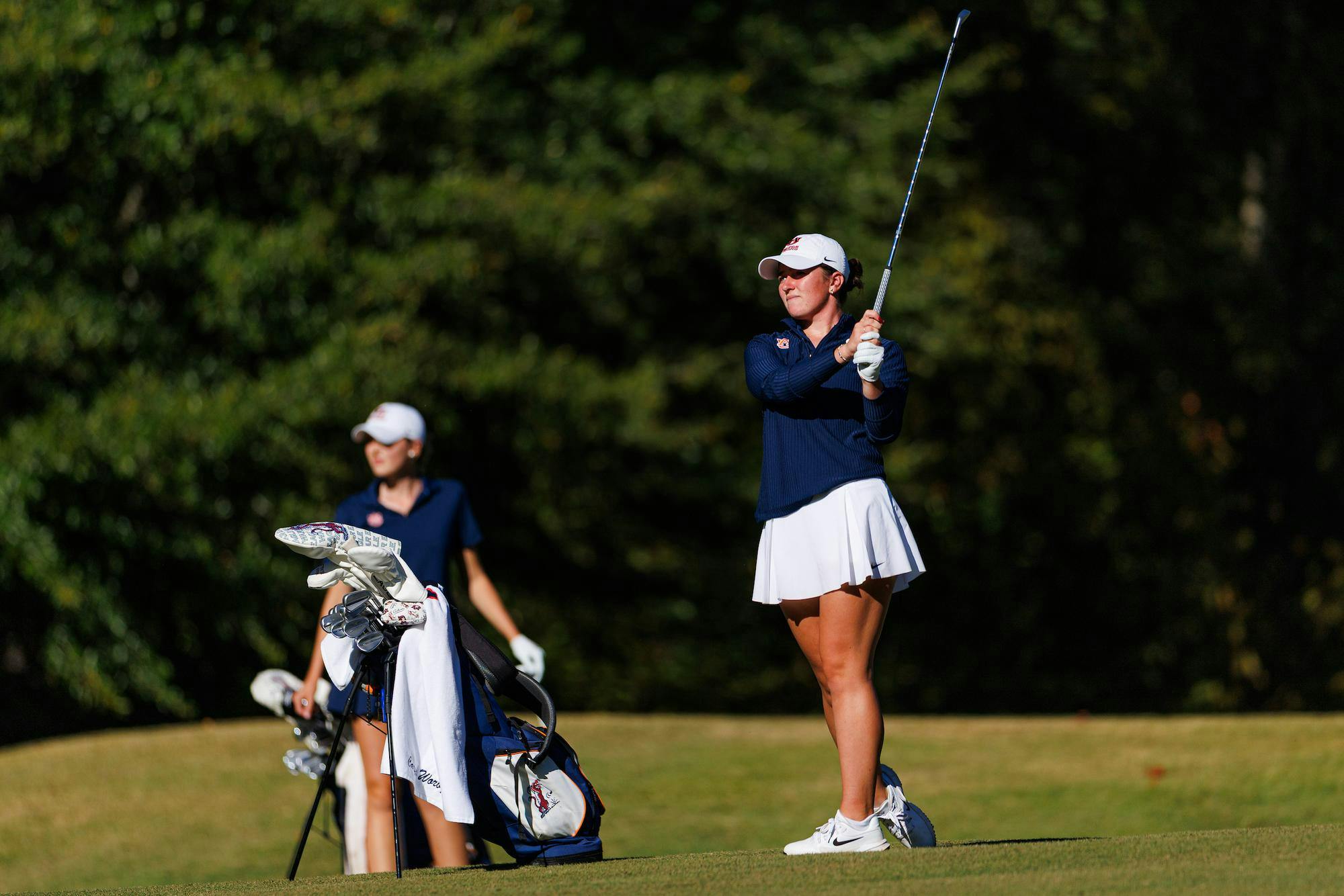 A female golfer in a blue top and white skirt prepares to swing a club, while a second golfer stands nearby with a golf bag. Contributed by Auburn Athletics