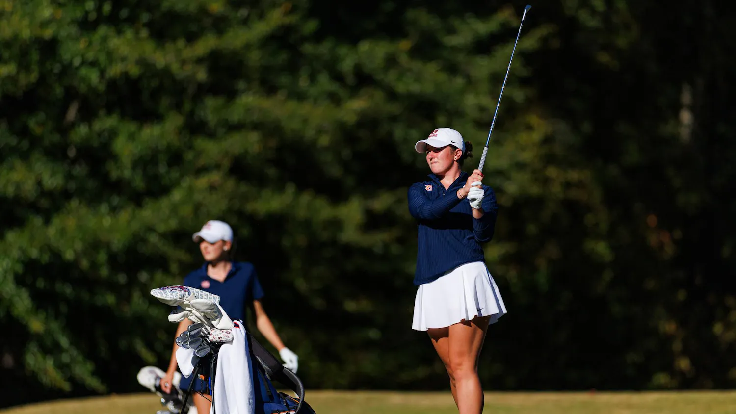 A female golfer in a blue top and white skirt prepares to swing a club, while a second golfer stands nearby with a golf bag. Contributed by Auburn Athletics