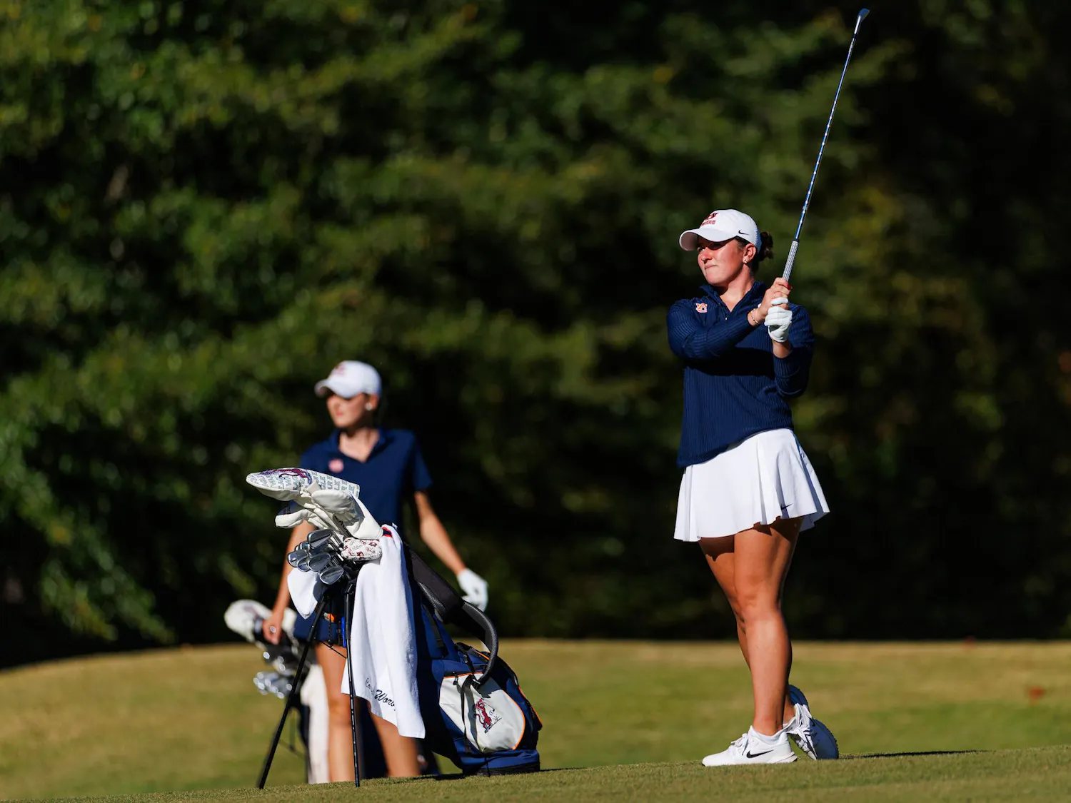 A female golfer in a blue top and white skirt prepares to swing a club, while a second golfer stands nearby with a golf bag. Contributed by Auburn Athletics