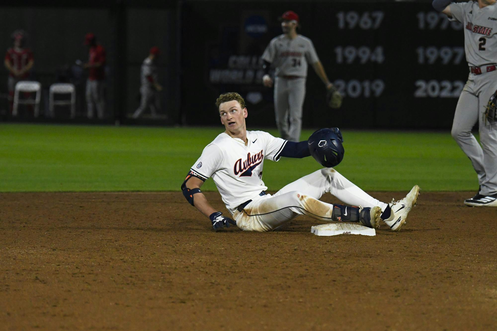 A baseball player in a white uniform sits on the ground near a base, looking back at other players in the background.