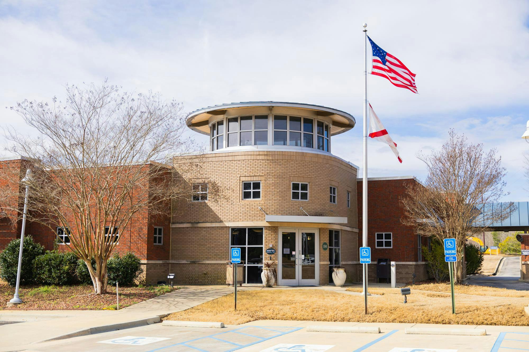 A modern brick building features a circular entrance, two flags on poles, and a landscaped parking area.