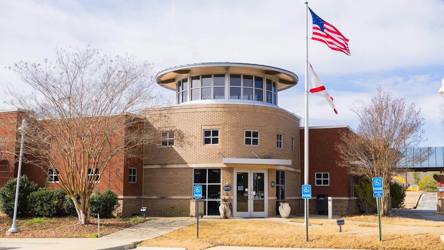 A modern brick building features a circular entrance, two flags on poles, and a landscaped parking area.