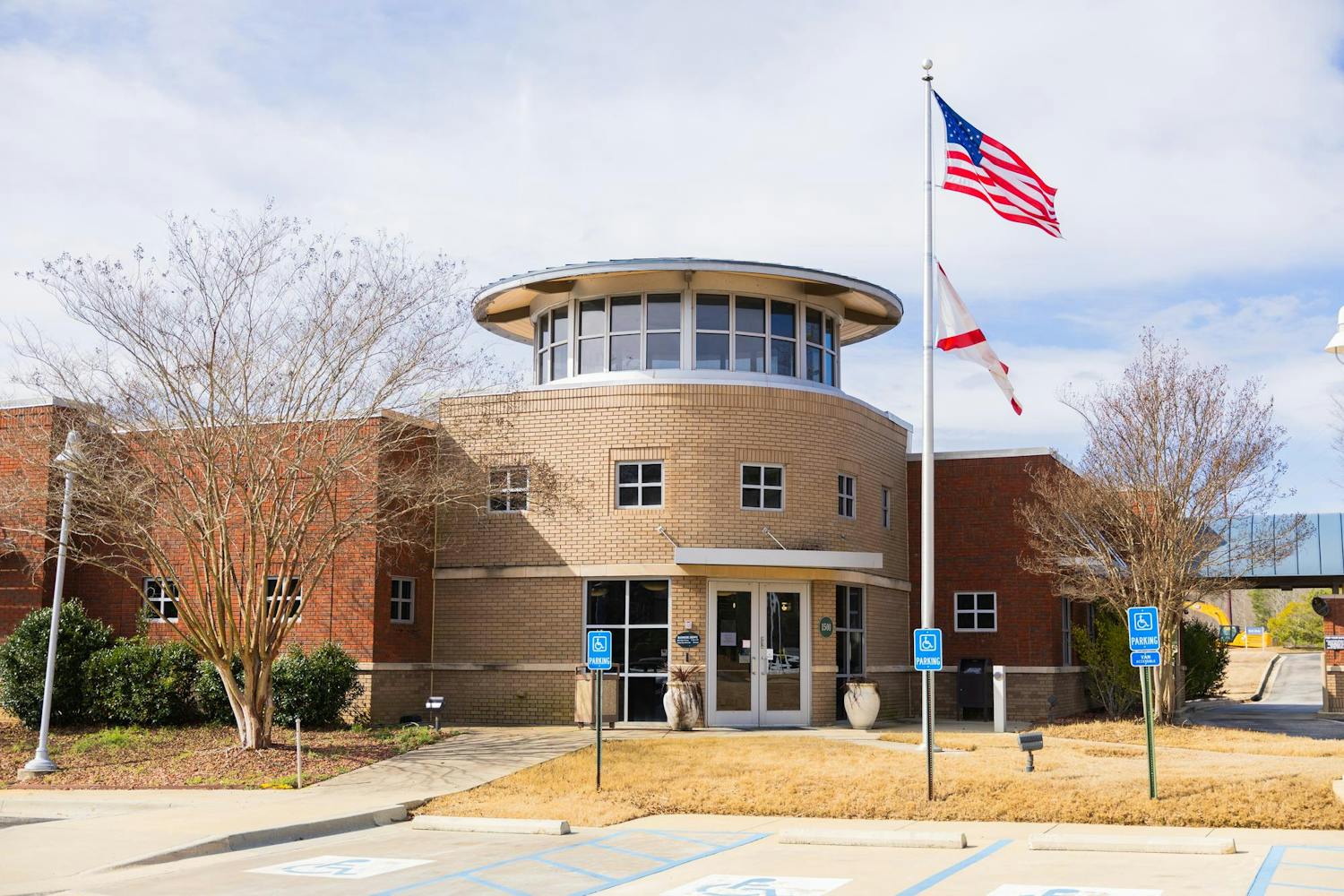 A modern brick building features a circular entrance, two flags on poles, and a landscaped parking area.
