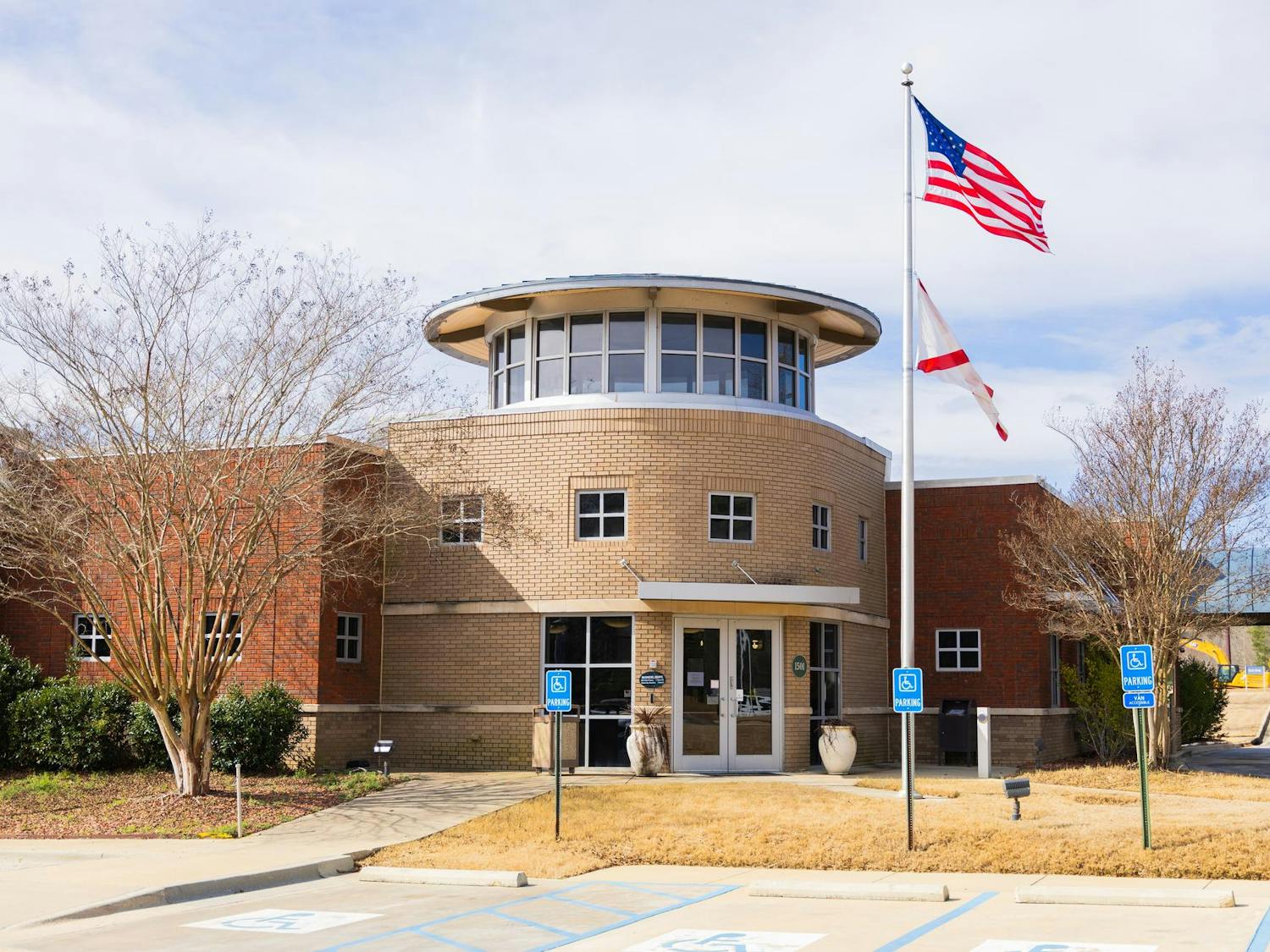 A modern brick building features a circular entrance, two flags on poles, and a landscaped parking area.
