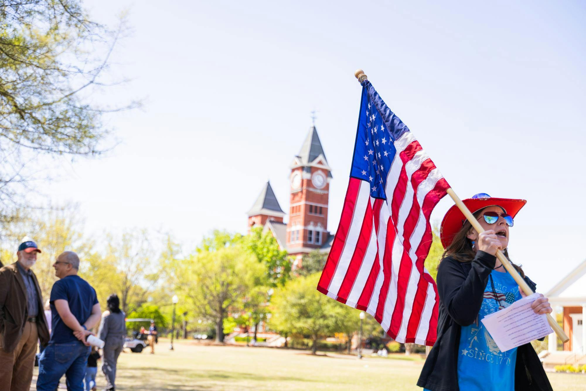 A woman in a red hat waves an American flag while standing near a clock tower and talking to people around her.