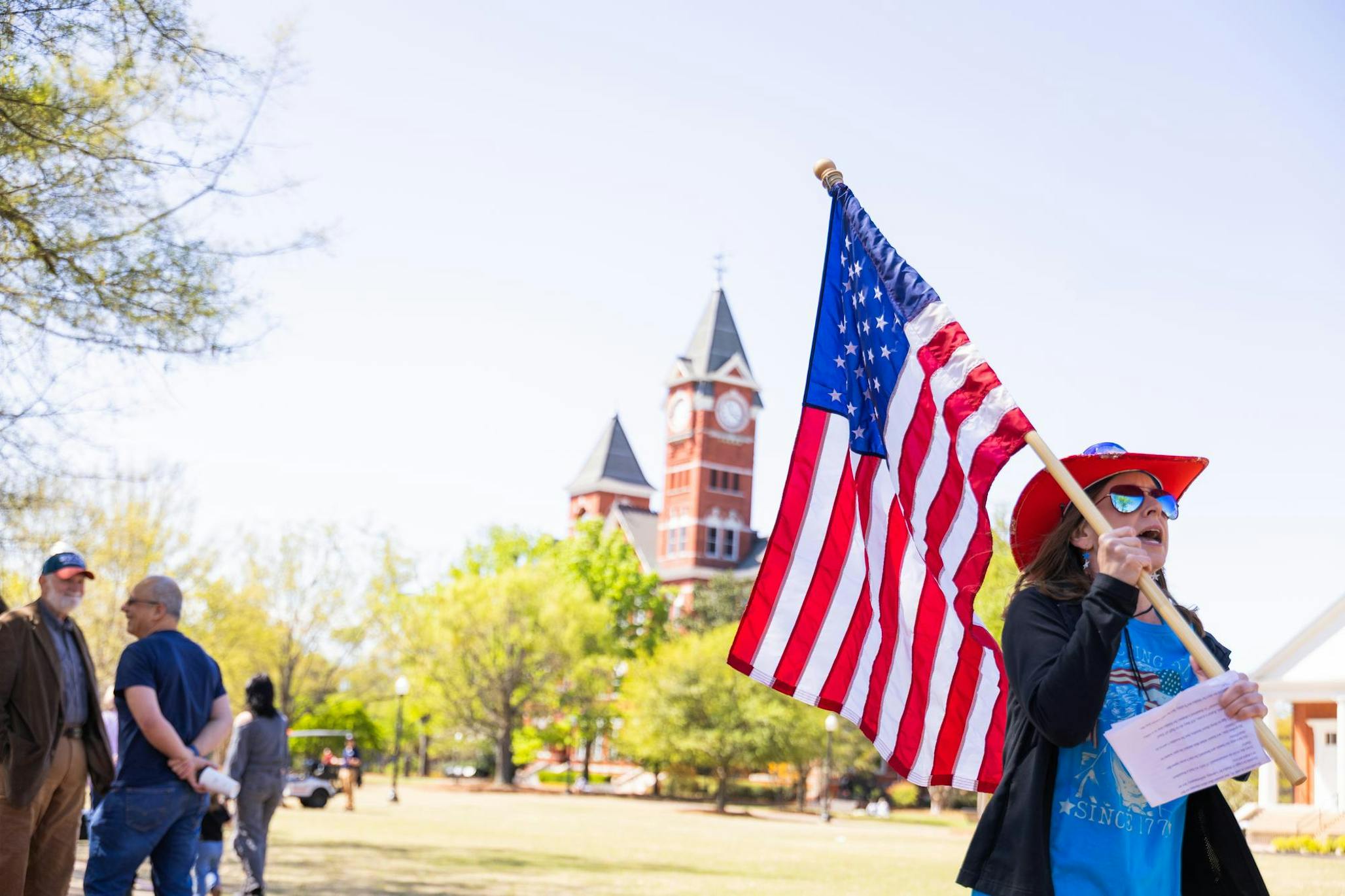 A woman in a red hat waves an American flag while standing near a clock tower and talking to people around her.