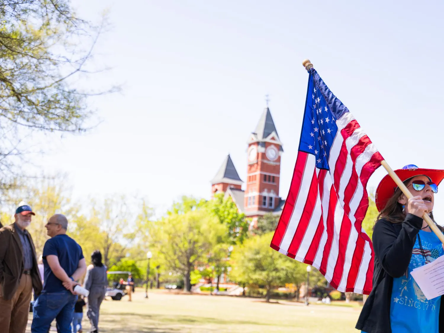 A woman in a red hat waves an American flag while standing near a clock tower and talking to people around her.