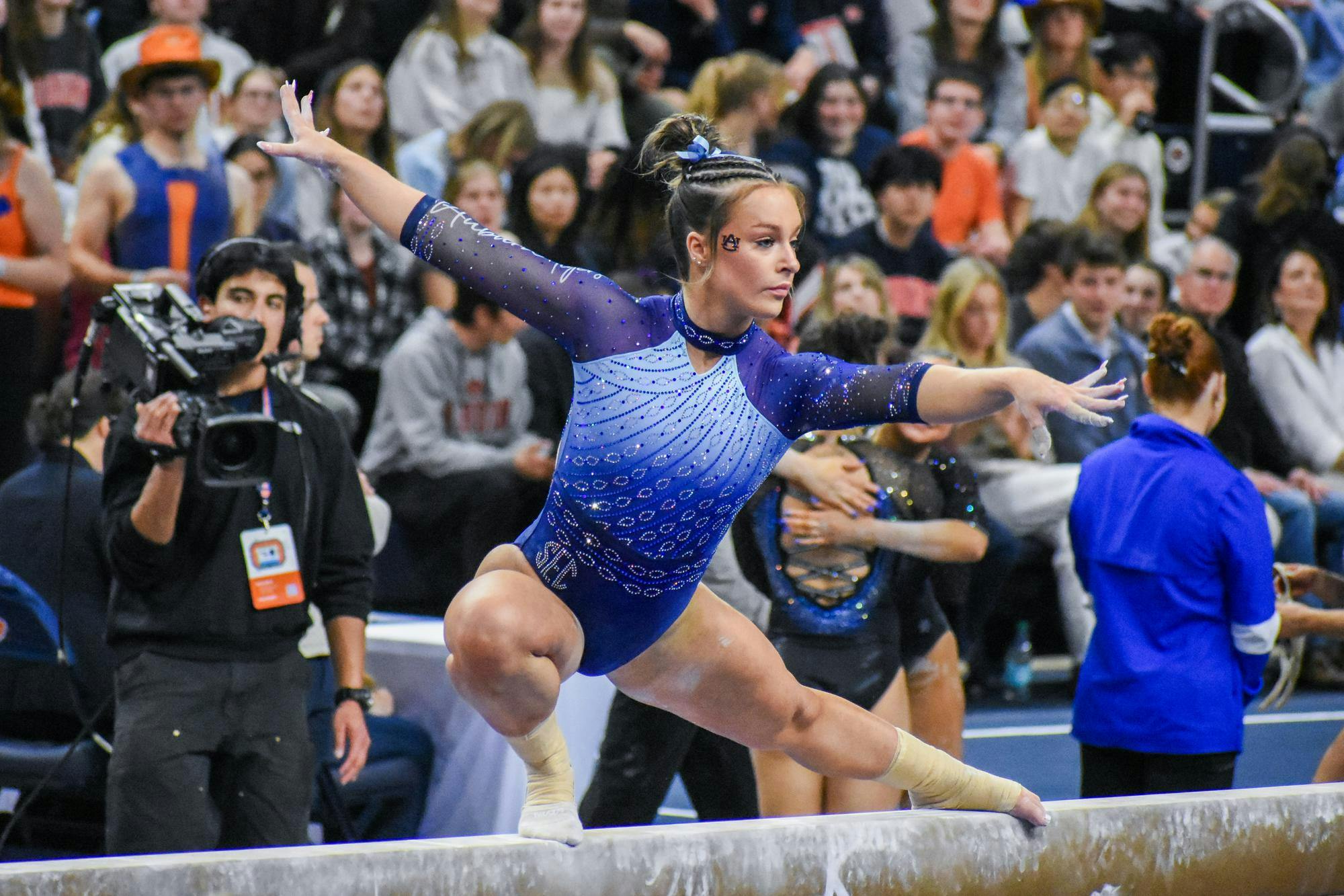 A gymnast in a blue and silver leotard performs on a balance beam while an audience watches, capturing the moment with a camera.