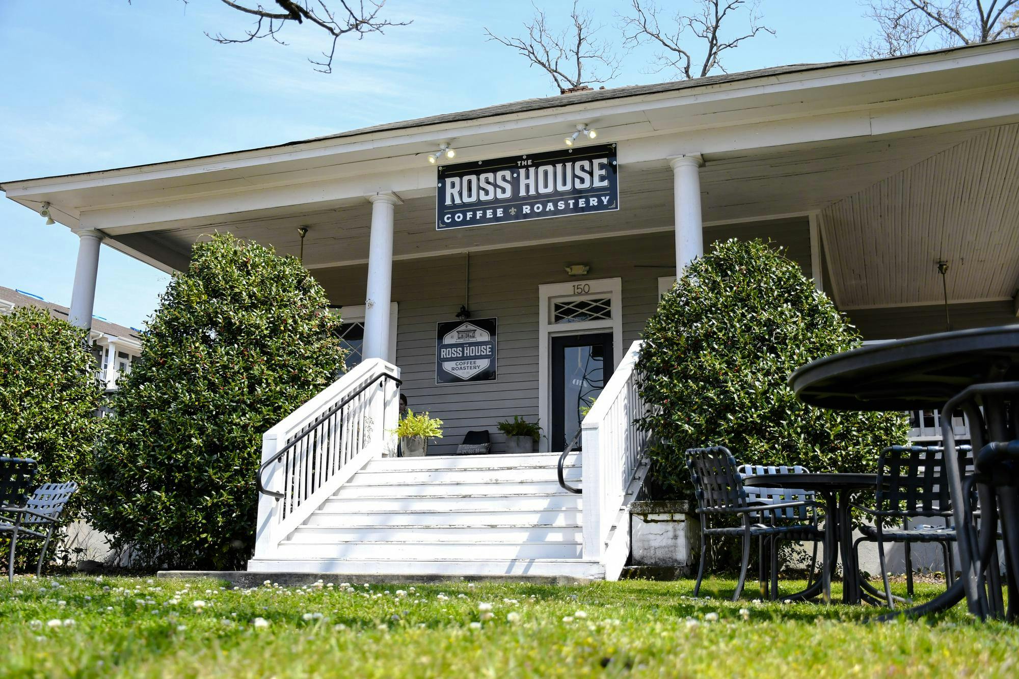 A spacious café with a large porch, white steps, green shrubs, and outdoor seating on a grassy area.