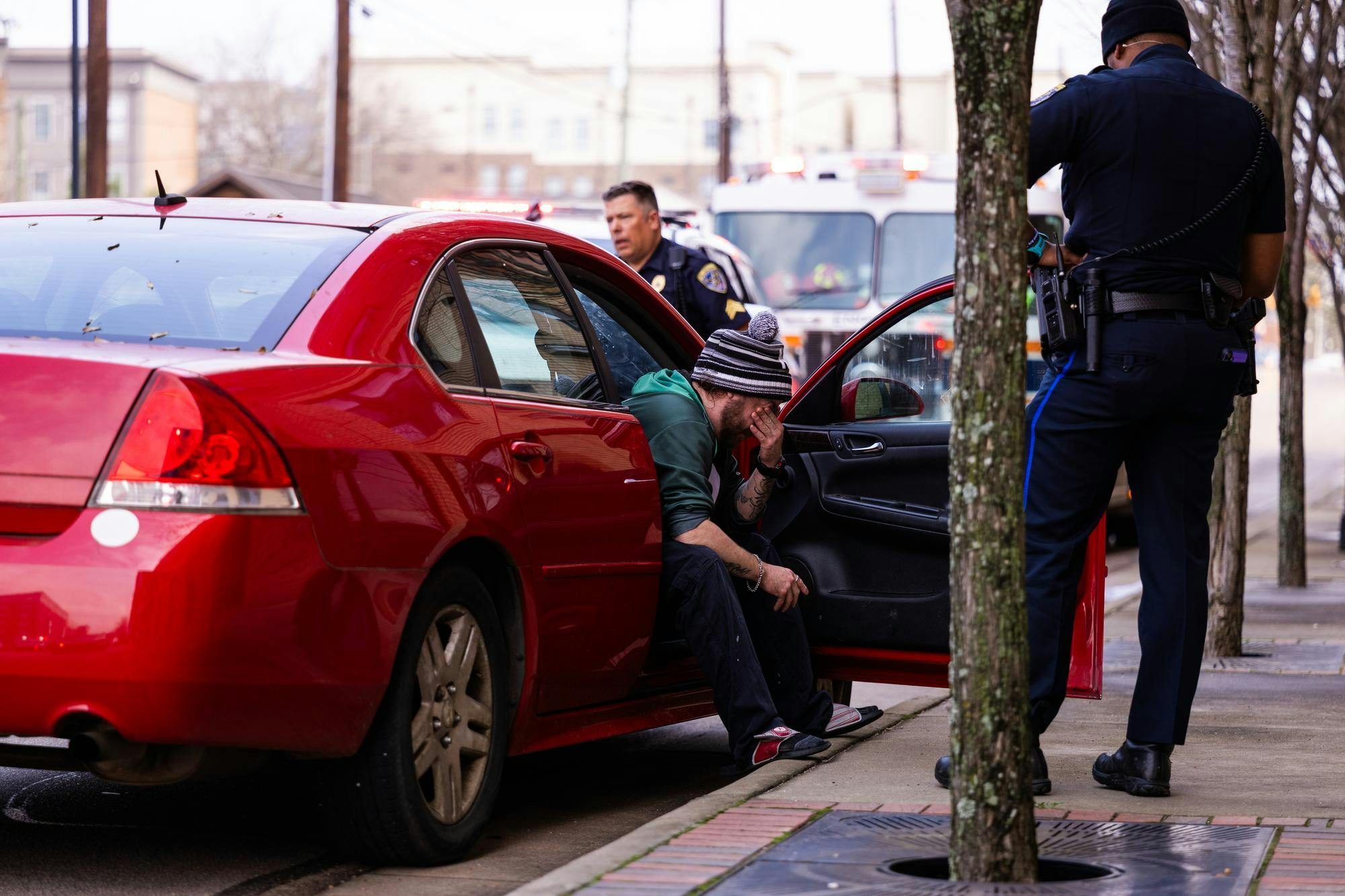 A man wearing a striped beanie sits distressed in a red car while a police officer stands nearby.