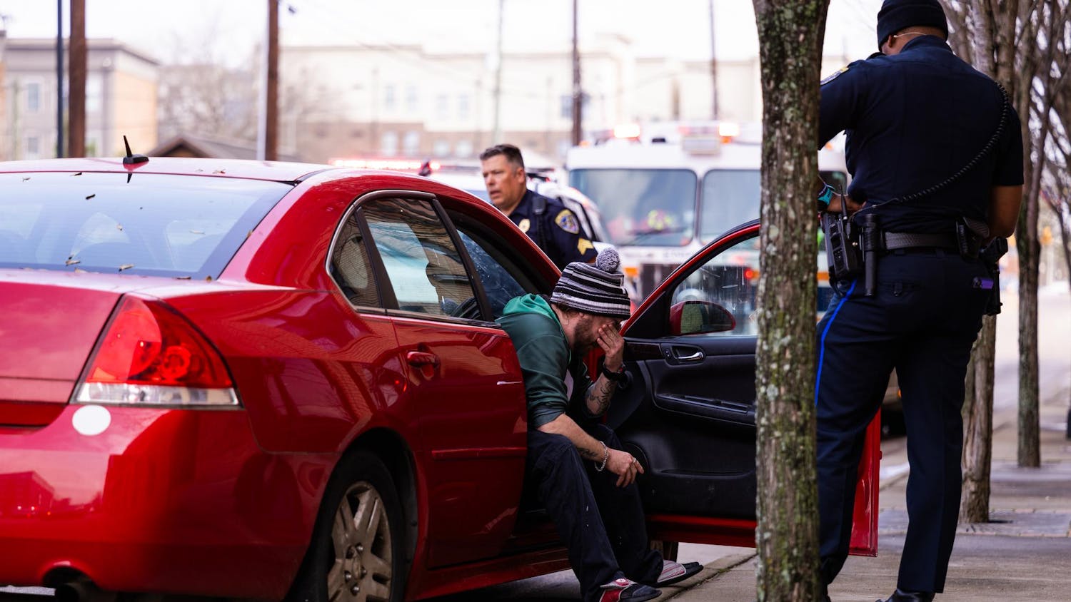 A man wearing a striped beanie sits distressed in a red car while a police officer stands nearby.