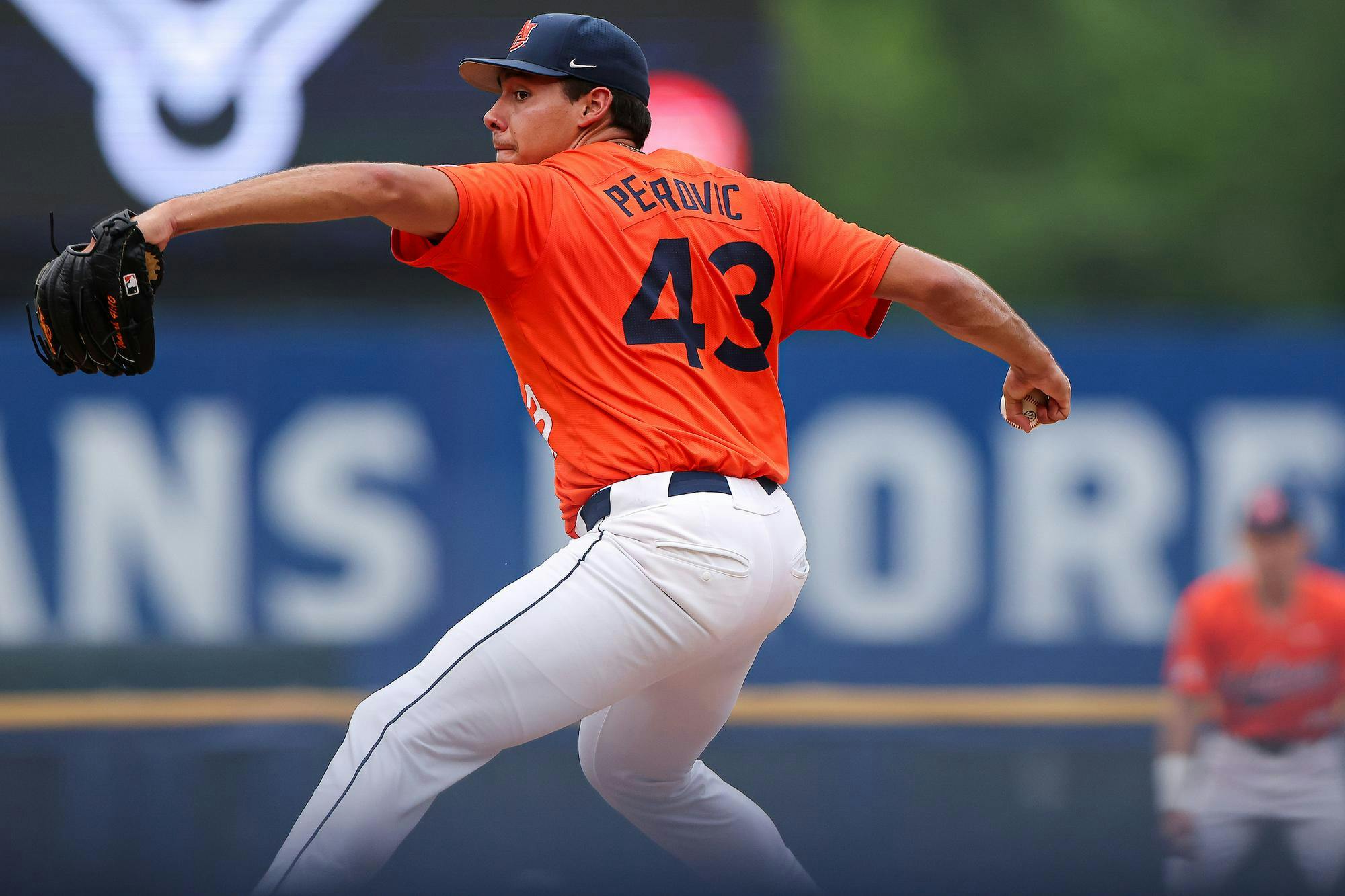 A baseball player in an orange uniform prepares to pitch on a field, with another player visible in the background.