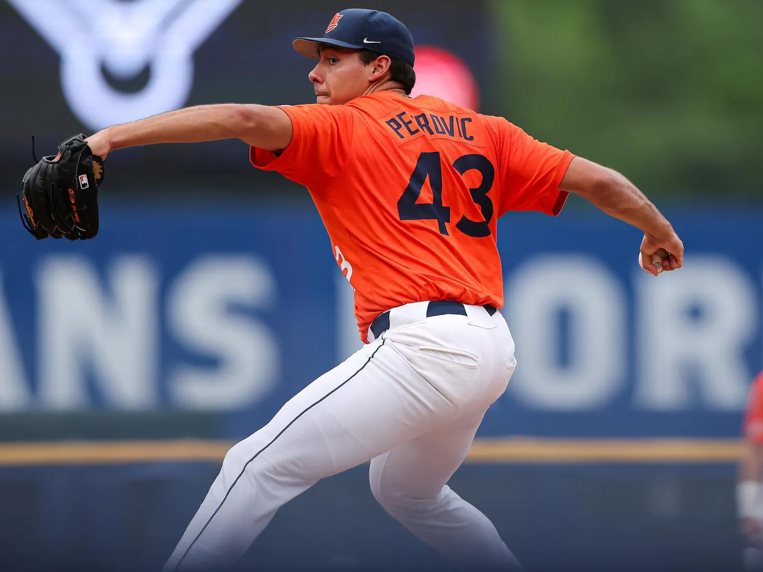 A baseball player in an orange uniform prepares to pitch on a field, with another player visible in the background.