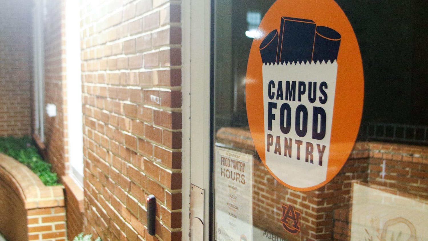 The Auburn Campus Food Pantry's glass door, which features an orange circular sign that reads "CAMPUS FOOD PANTRY," and information about food pantry hours is visible below.