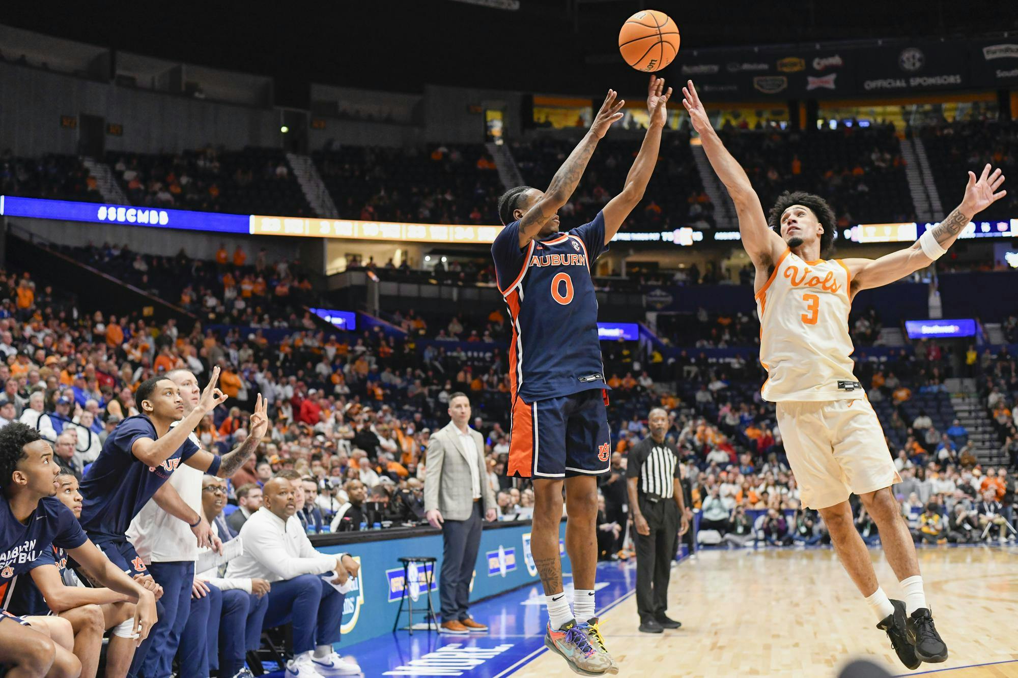 A player in an orange jersey shoots a basketball while another player in a dark blue jersey attempts to block the shot.