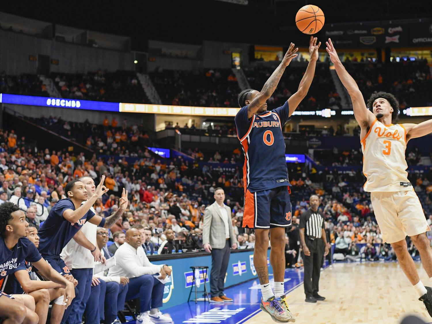 A player in an orange jersey shoots a basketball while another player in a dark blue jersey attempts to block the shot.