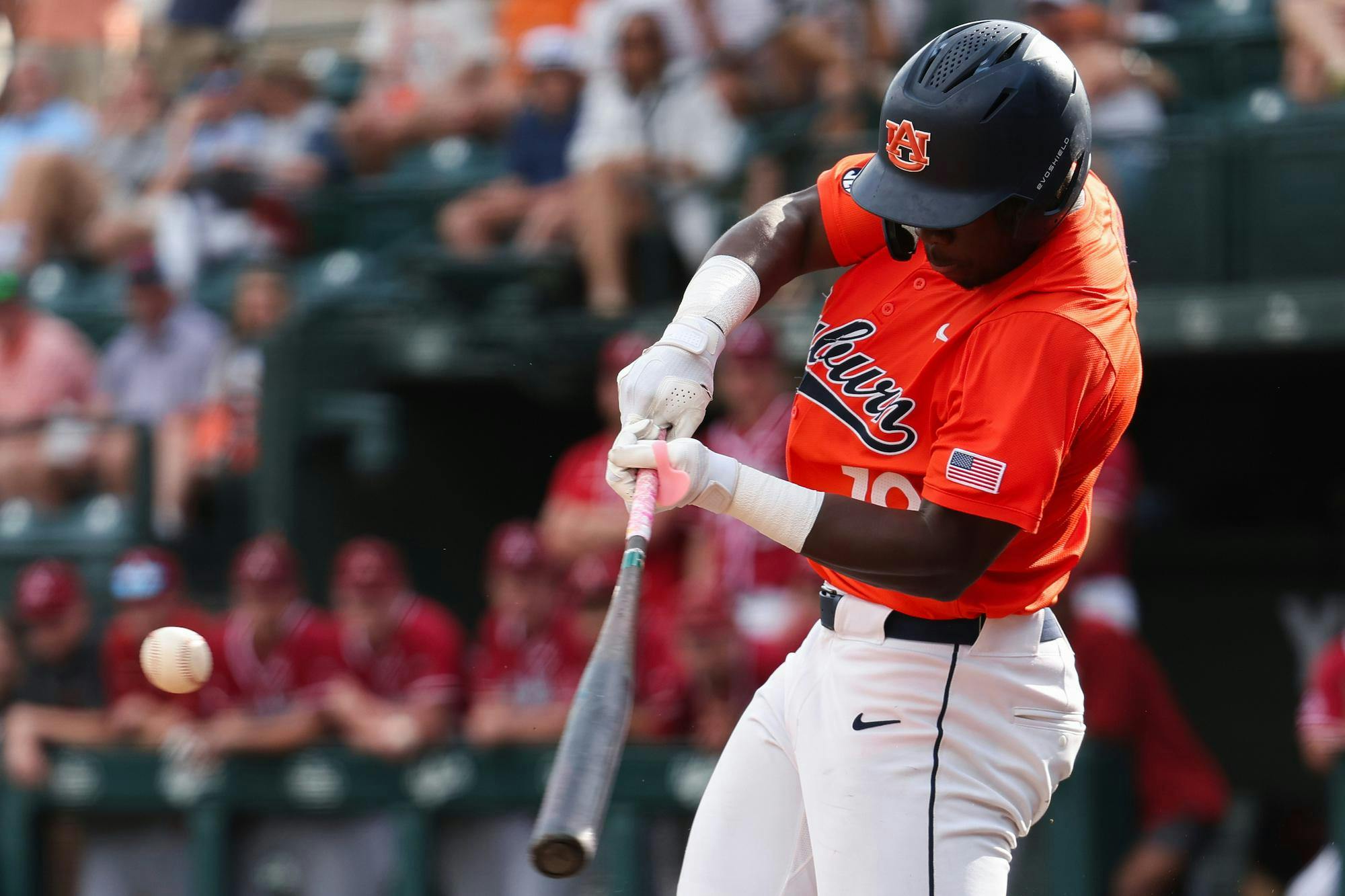 A baseball player in an orange uniform swings a bat at a baseball while spectators watch in the background.