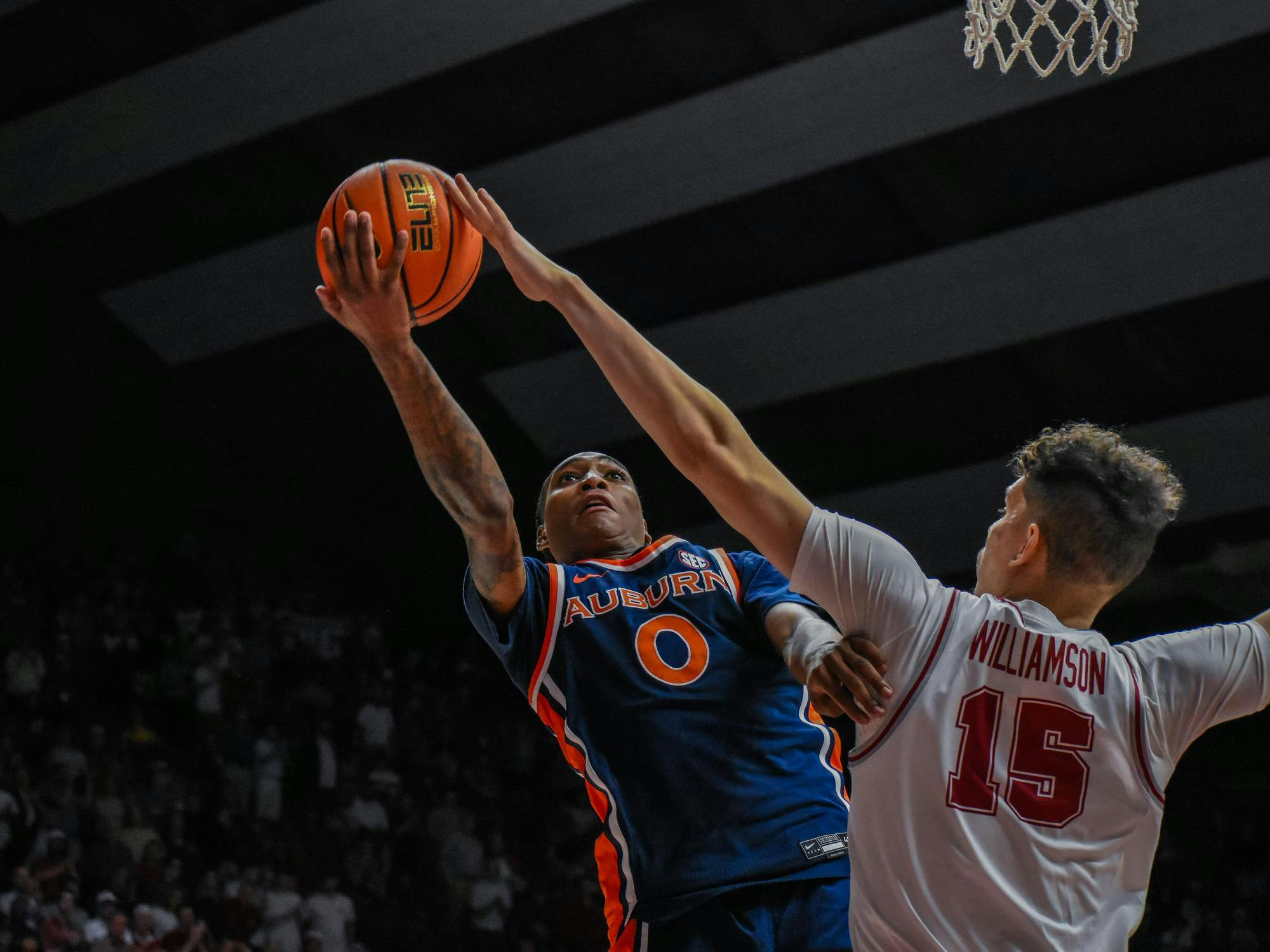 A player in an orange and blue uniform attempts a layup while being challenged by another player in a white uniform.