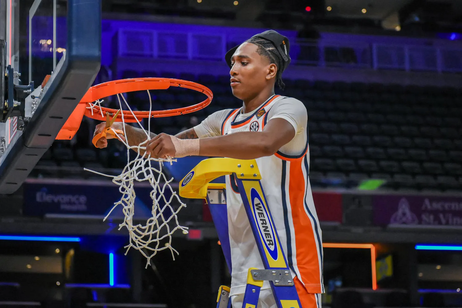 A young athlete in a basketball uniform is cutting down a net from a hoop while standing on a ladder.