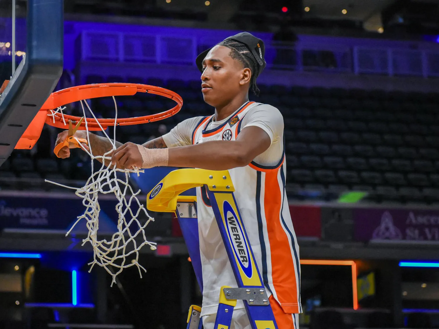 A young athlete in a basketball uniform is cutting down a net from a hoop while standing on a ladder.
