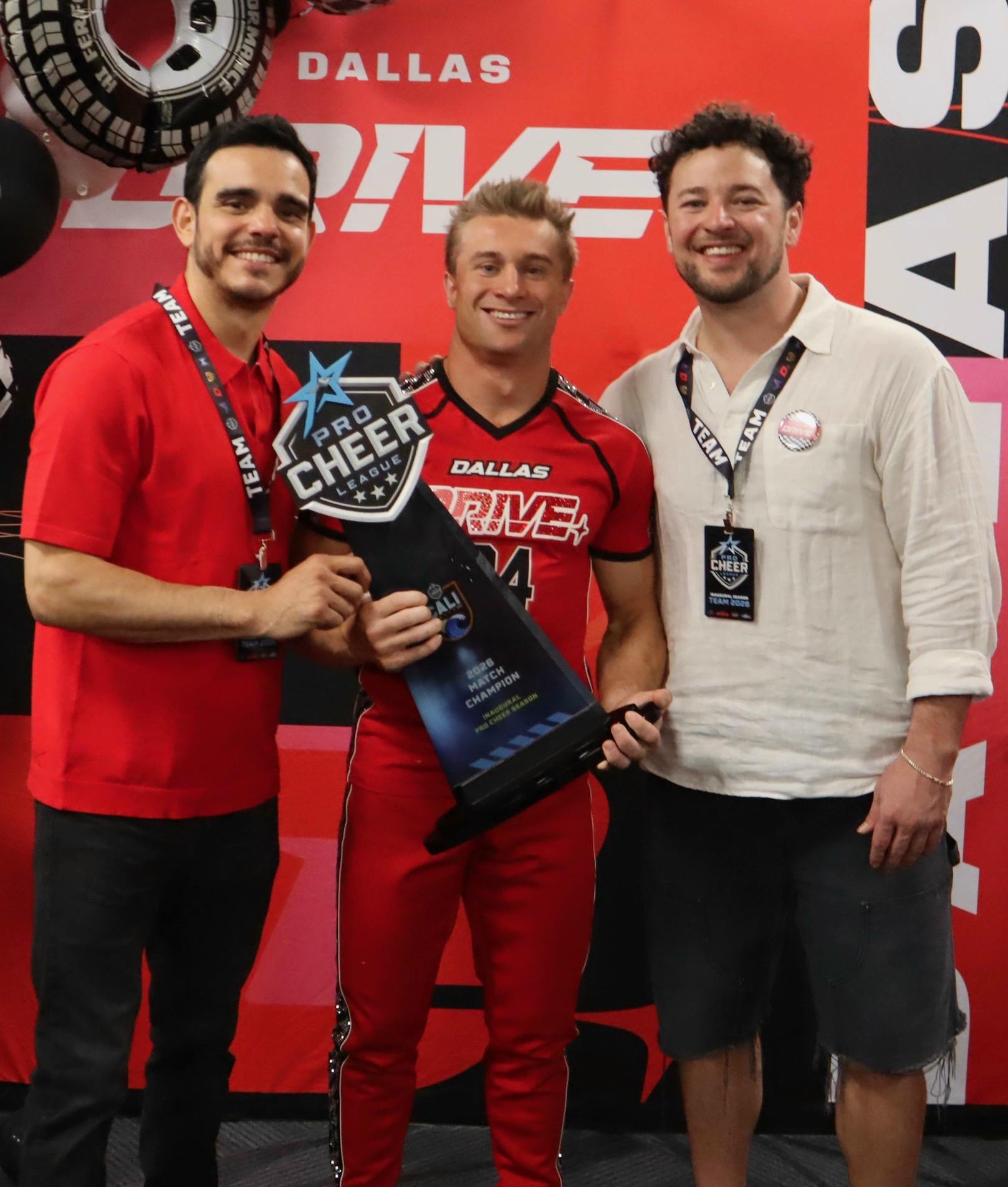 Three men stand together, smiling and posing with a trophy in front of a colorful backdrop featuring the text "DALLAS DRIVE."