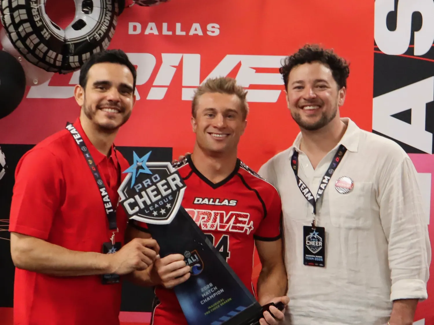 Three men stand together, smiling and posing with a trophy in front of a colorful backdrop featuring the text "DALLAS DRIVE."