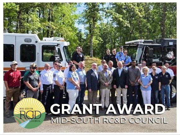 Opelika Fire Department stand together outdoors in a forested area with two emergency vehicles. Contributed by Mid South RC&D Council.