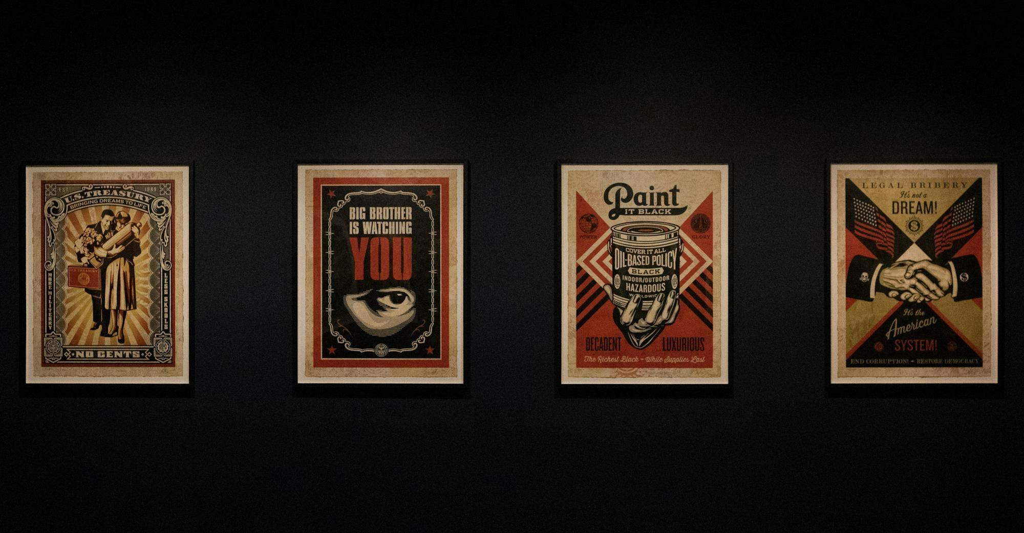 Four framed posters on a black wall feature bold text and imagery related to themes of capitalism, surveillance, and corruption.