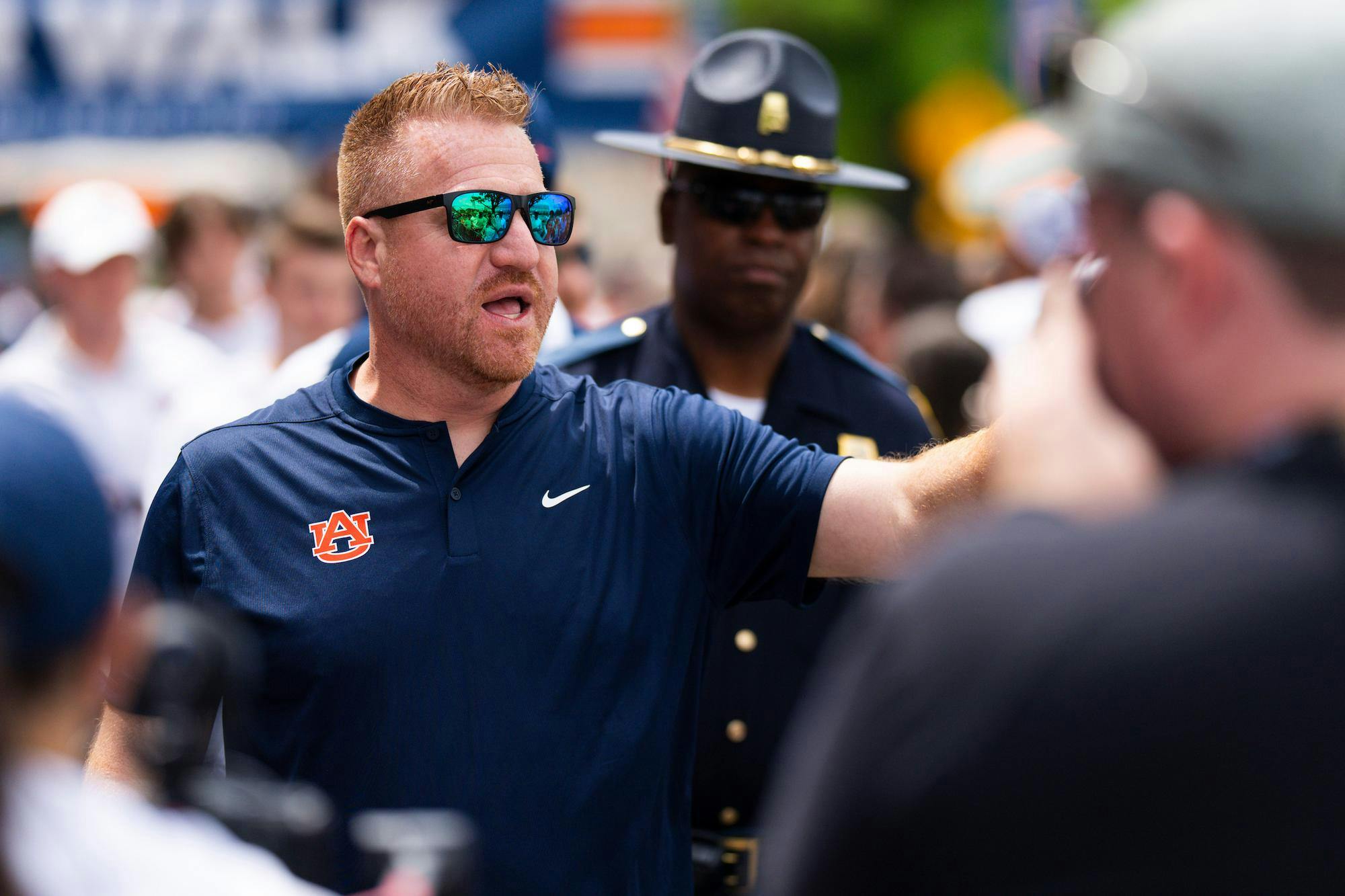 A man in a navy polo shirt with an Auburn logo gestures expressively while wearing sunglasses, surrounded by a crowd and a police officer.