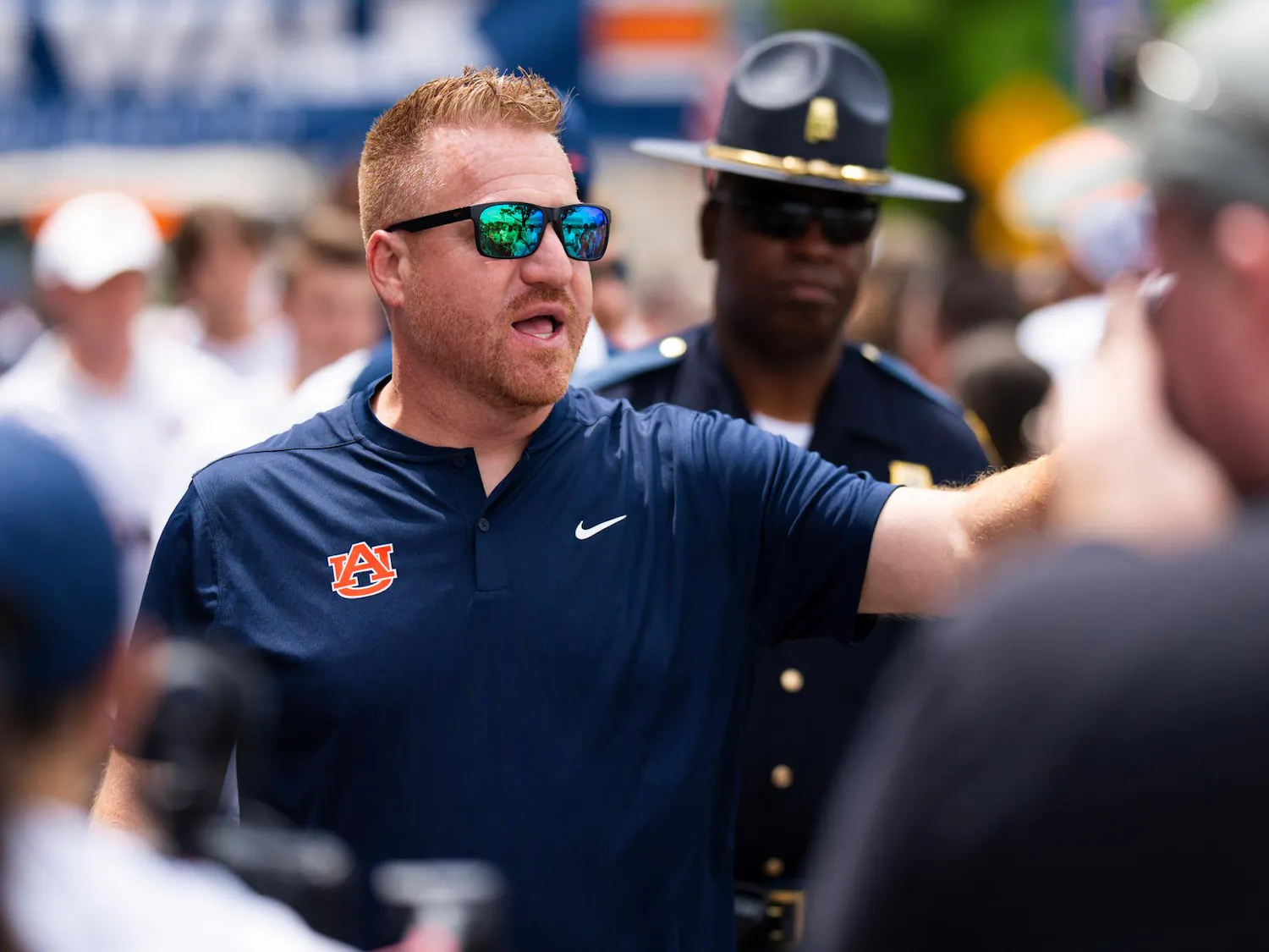 A man in a navy polo shirt with an Auburn logo gestures expressively while wearing sunglasses, surrounded by a crowd and a police officer.