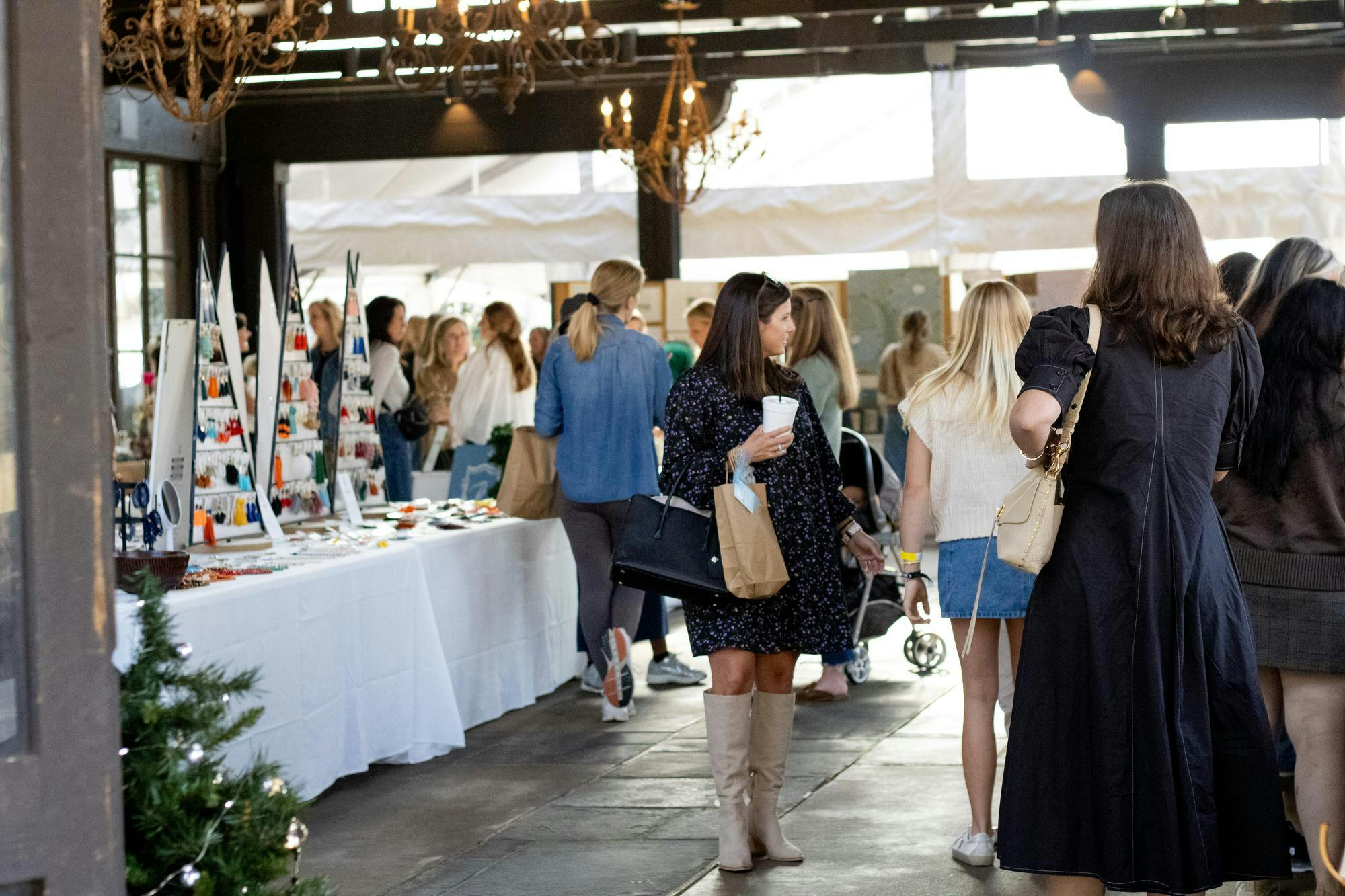 A bustling indoor market features shoppers browsing handmade jewelry displays and vintage goods, with a festive atmosphere highlighted by chandeliers and decorations.