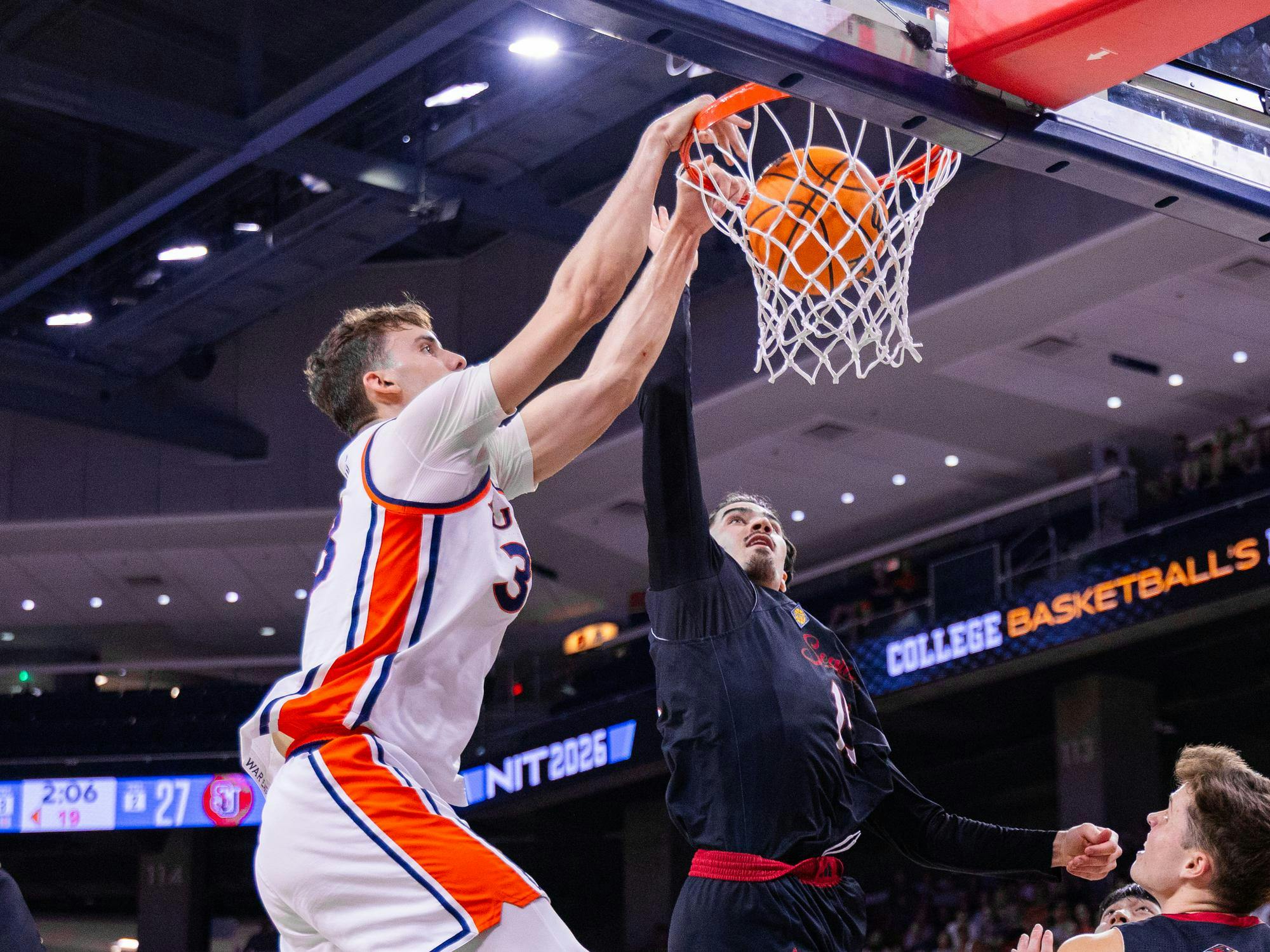 A player in a white jersey jumps to shoot a basketball while another player in black attempts to block the shot near the hoop.