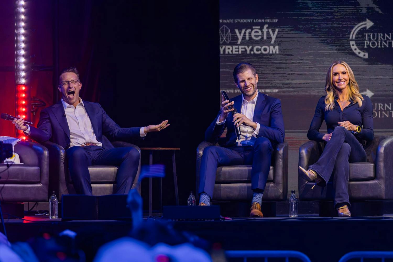 Three individuals sit on stage in armchairs, with one man animatedly speaking, while another holds a device and a woman smiles.