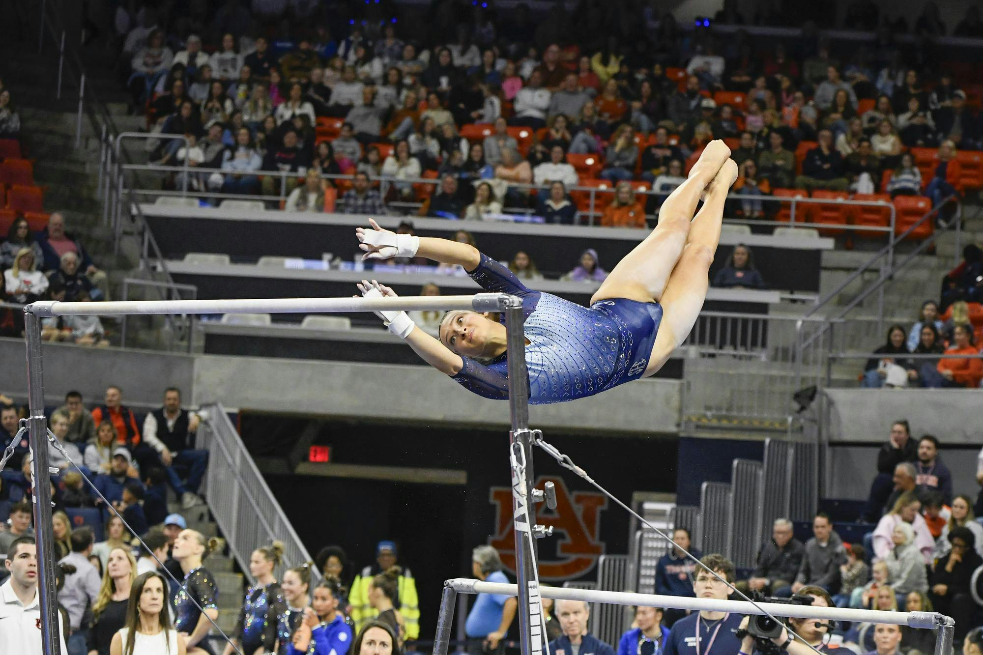 A gymnast in a blue outfit is performing an aerial move on uneven bars, while a crowd watches from the stands.