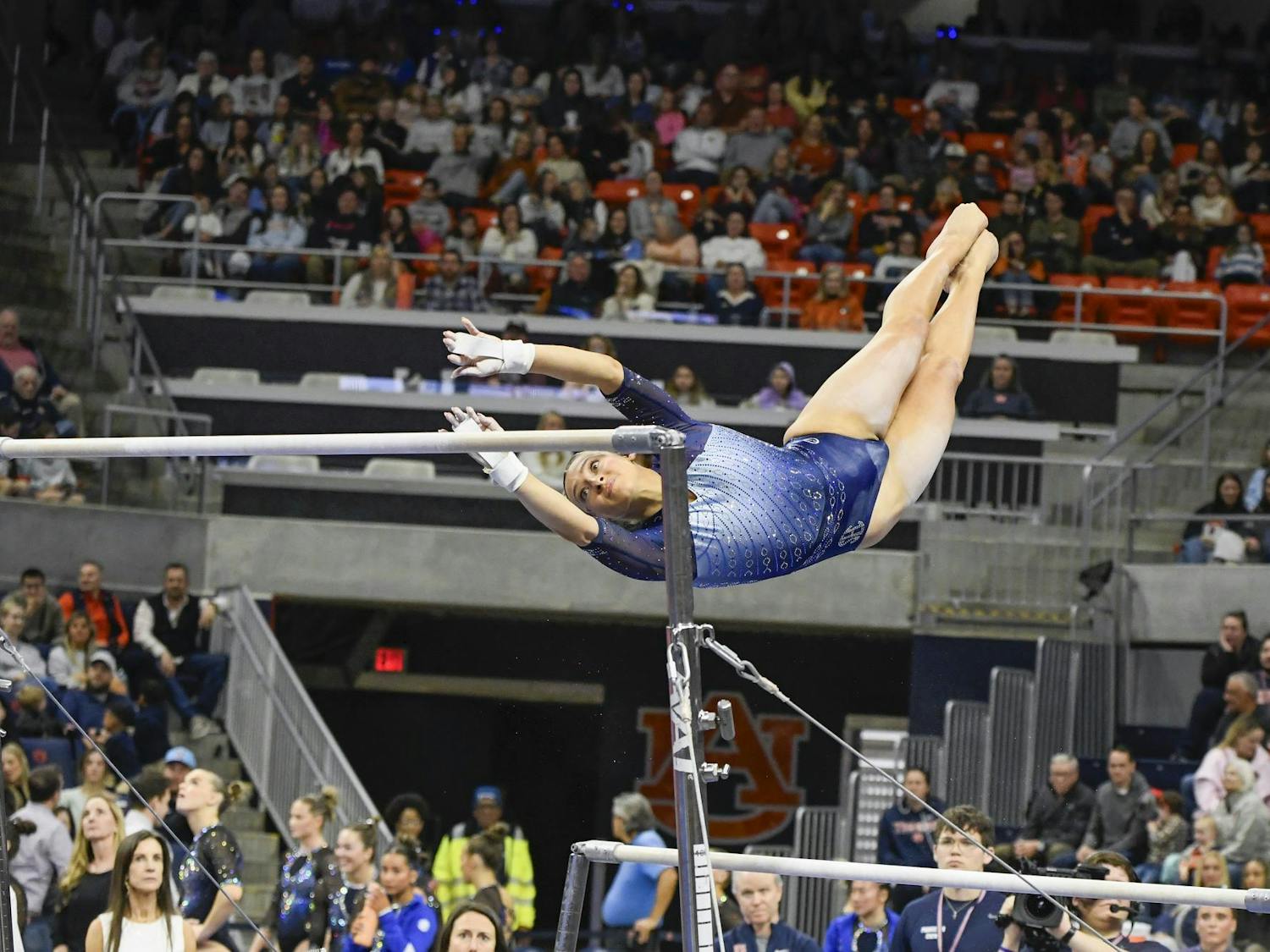 A gymnast in a blue outfit is performing an aerial move on uneven bars, while a crowd watches from the stands.