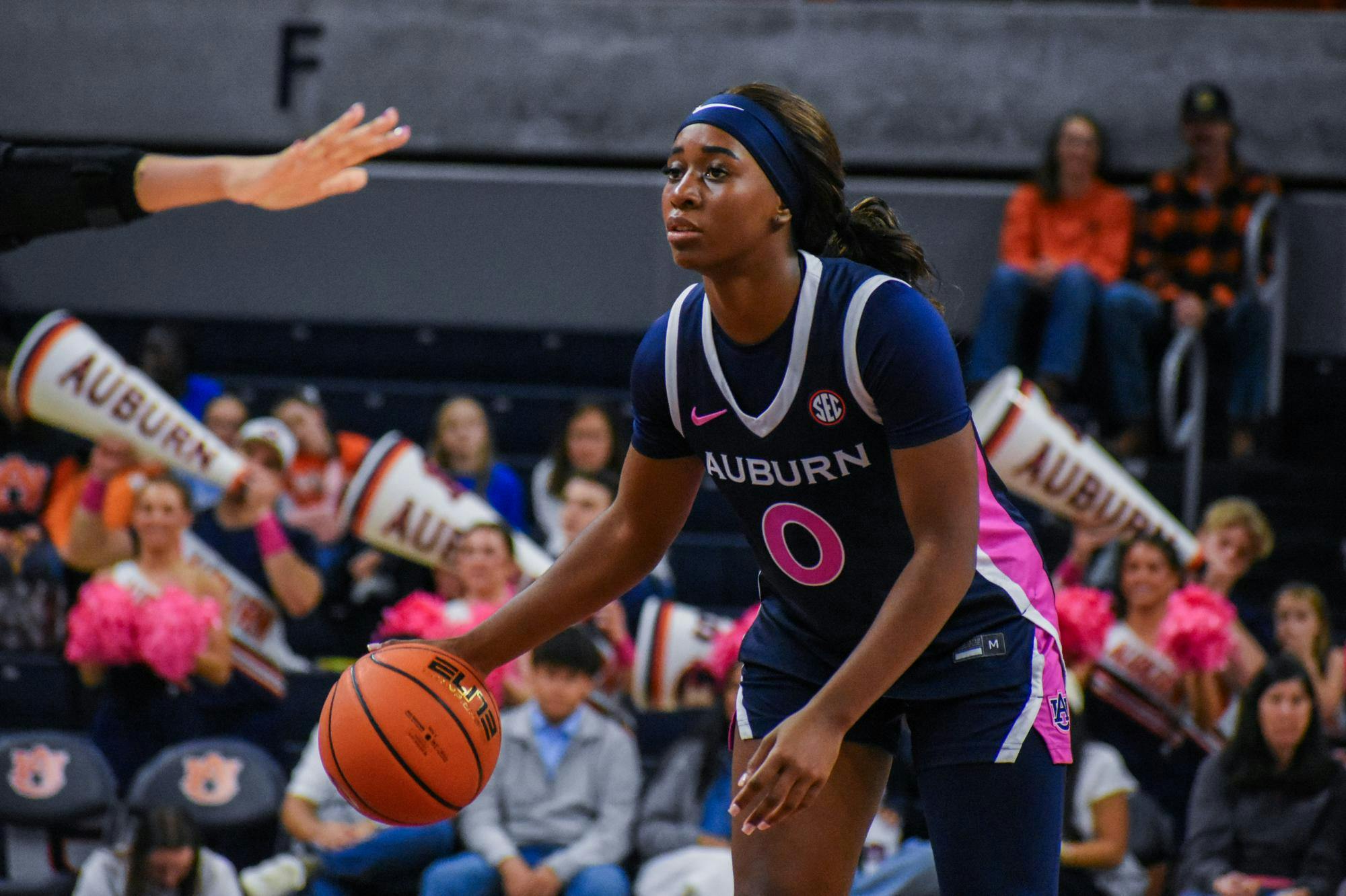 A basketball player wearing a dark uniform with "Auburn" written on it prepares to dribble a basketball amidst cheering fans holding pom-poms and megaphones.