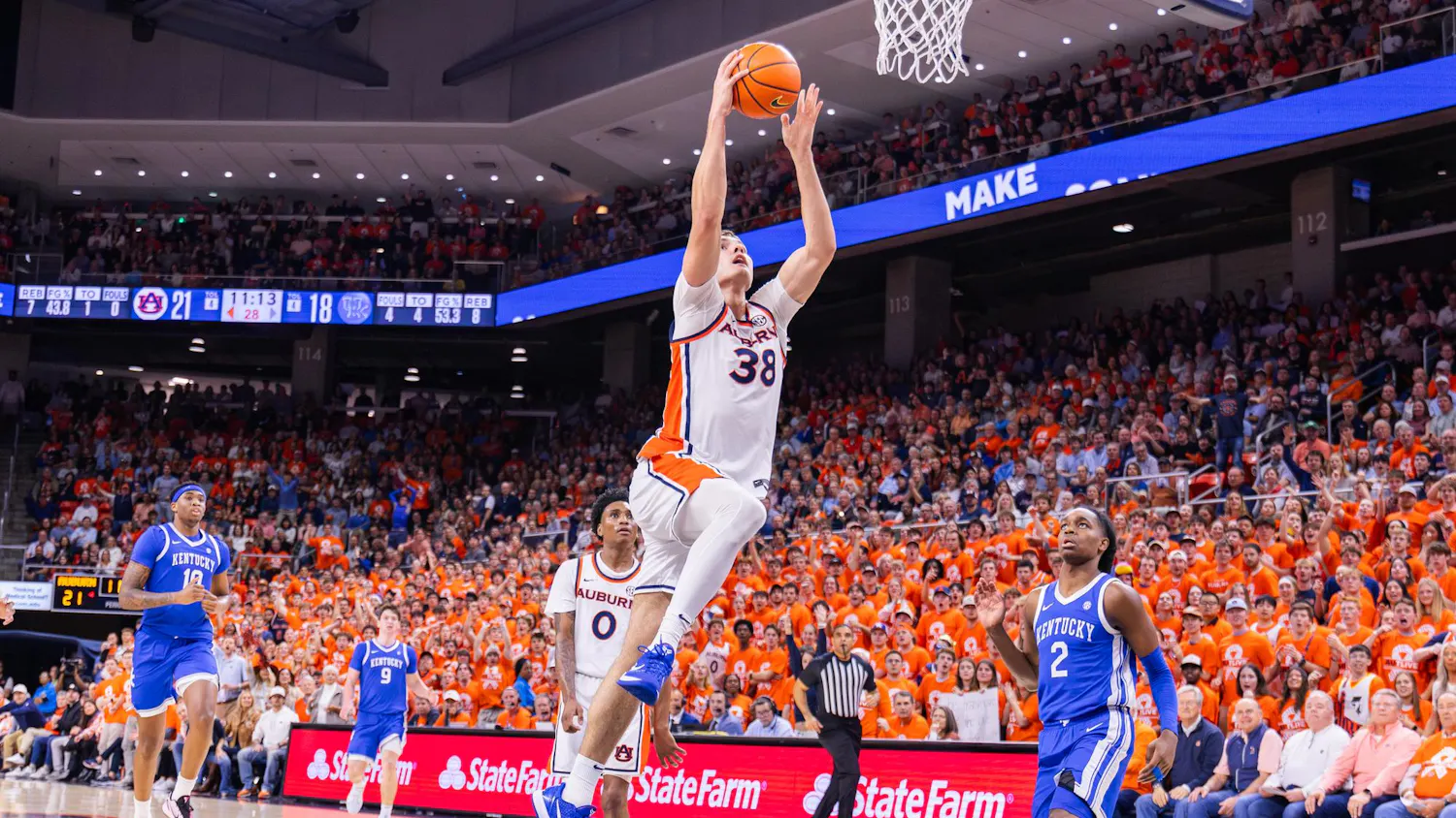 A player leaps toward the basketball hoop, while teammates and opponents watch, amidst a crowd dressed in orange.