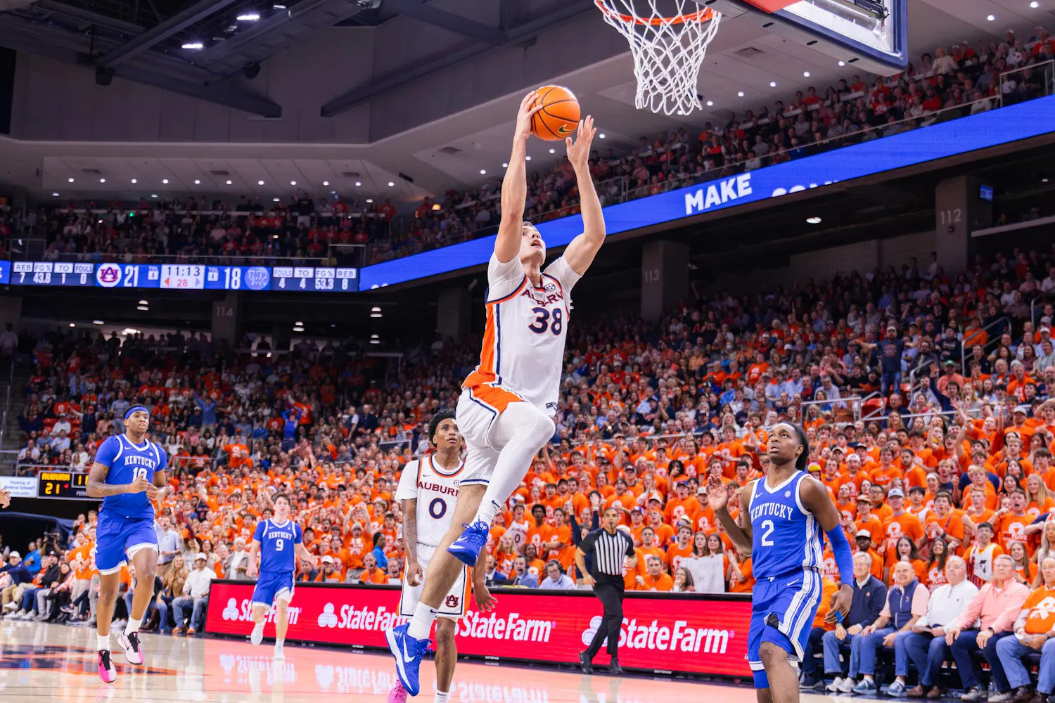 A player leaps toward the basketball hoop, while teammates and opponents watch, amidst a crowd dressed in orange.