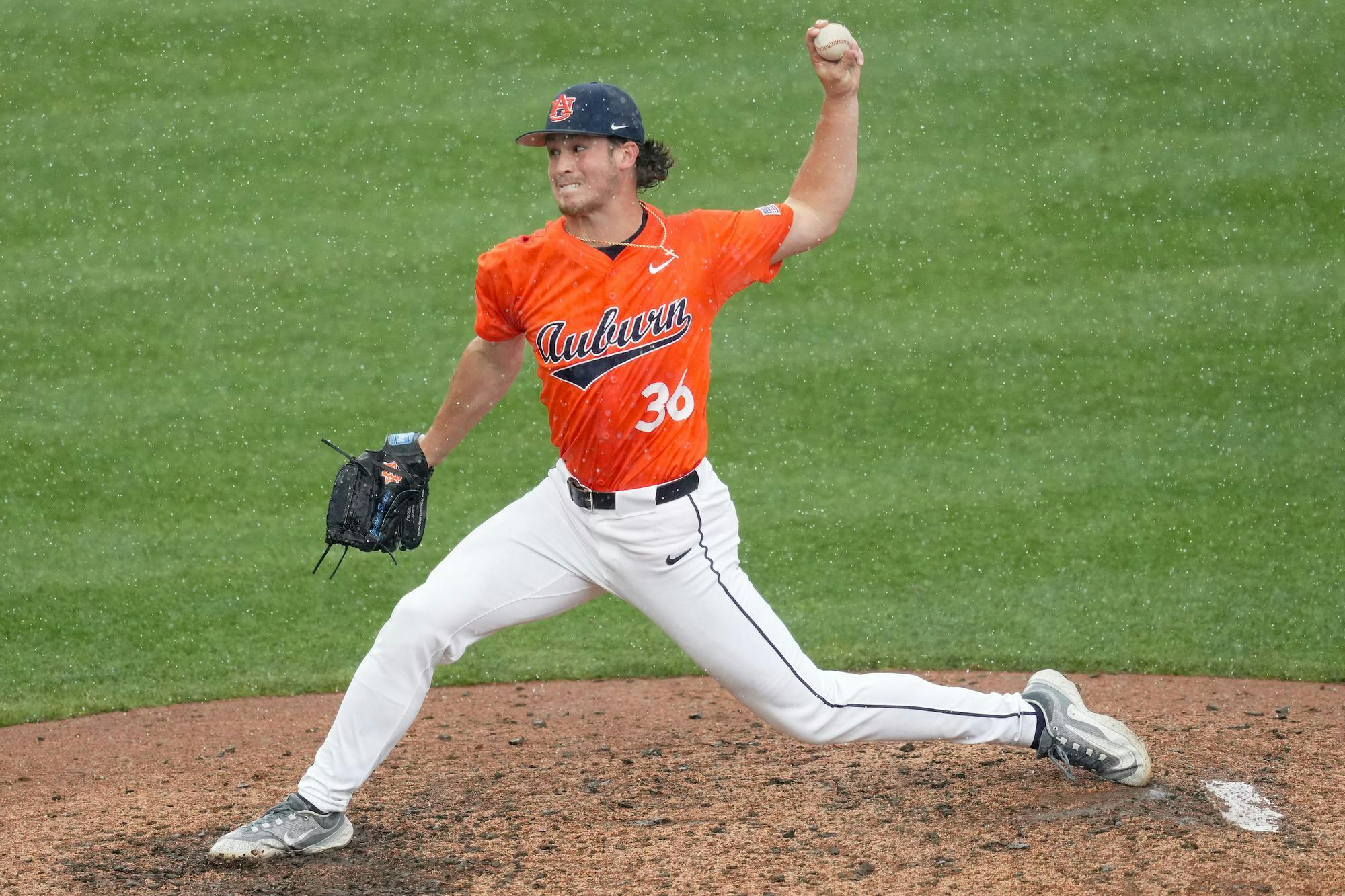 A baseball pitcher in an orange jersey throws a pitch on a raining field, with droplets visible around him.
