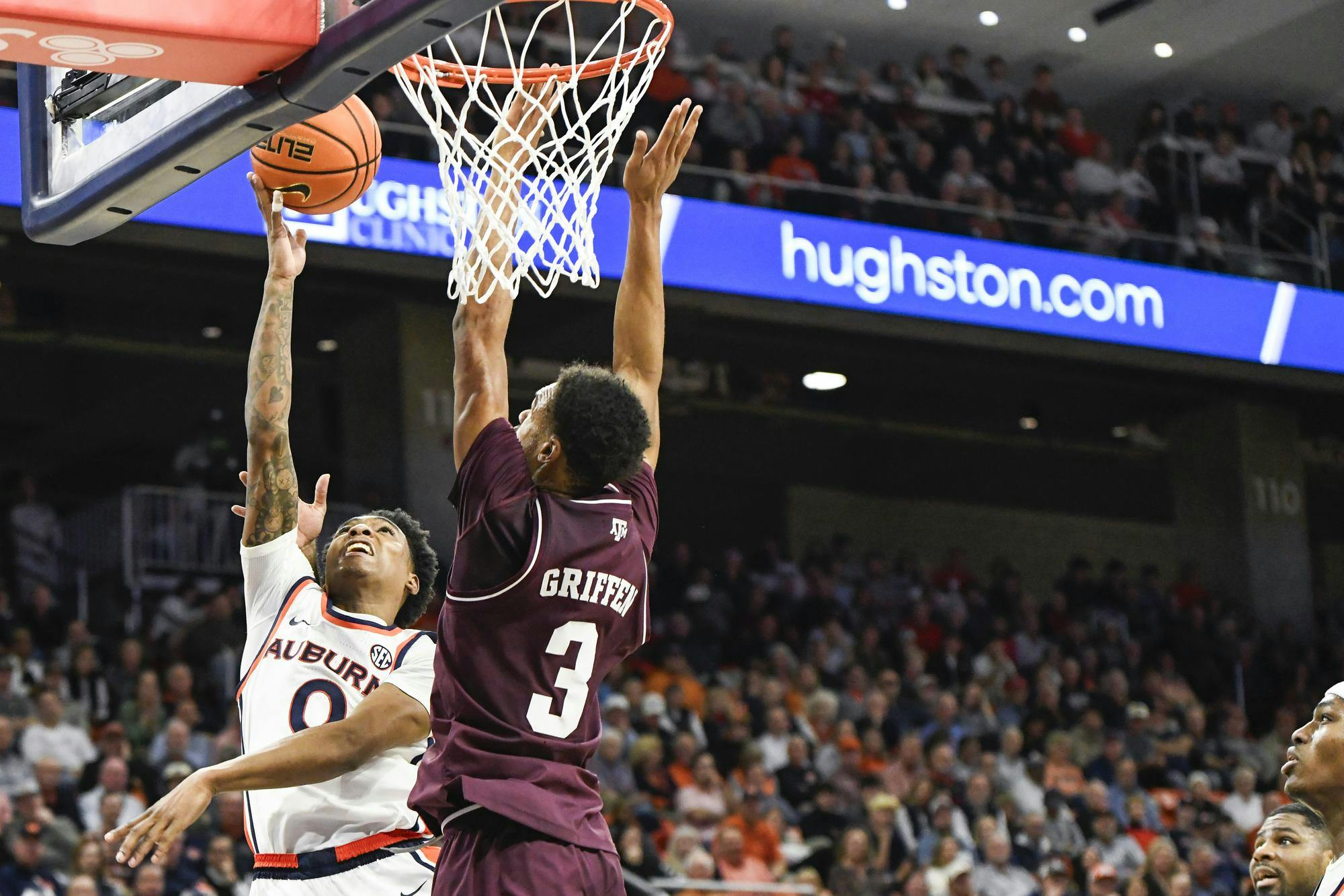 A basketball player in an orange and white uniform leaps for a shot while another player in maroon tries to block him.