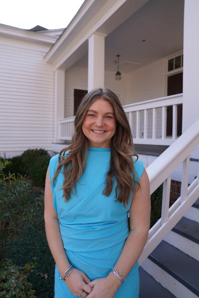 A woman in a blue dress stands outdoors, smiling with a white house and greenery in the background.
