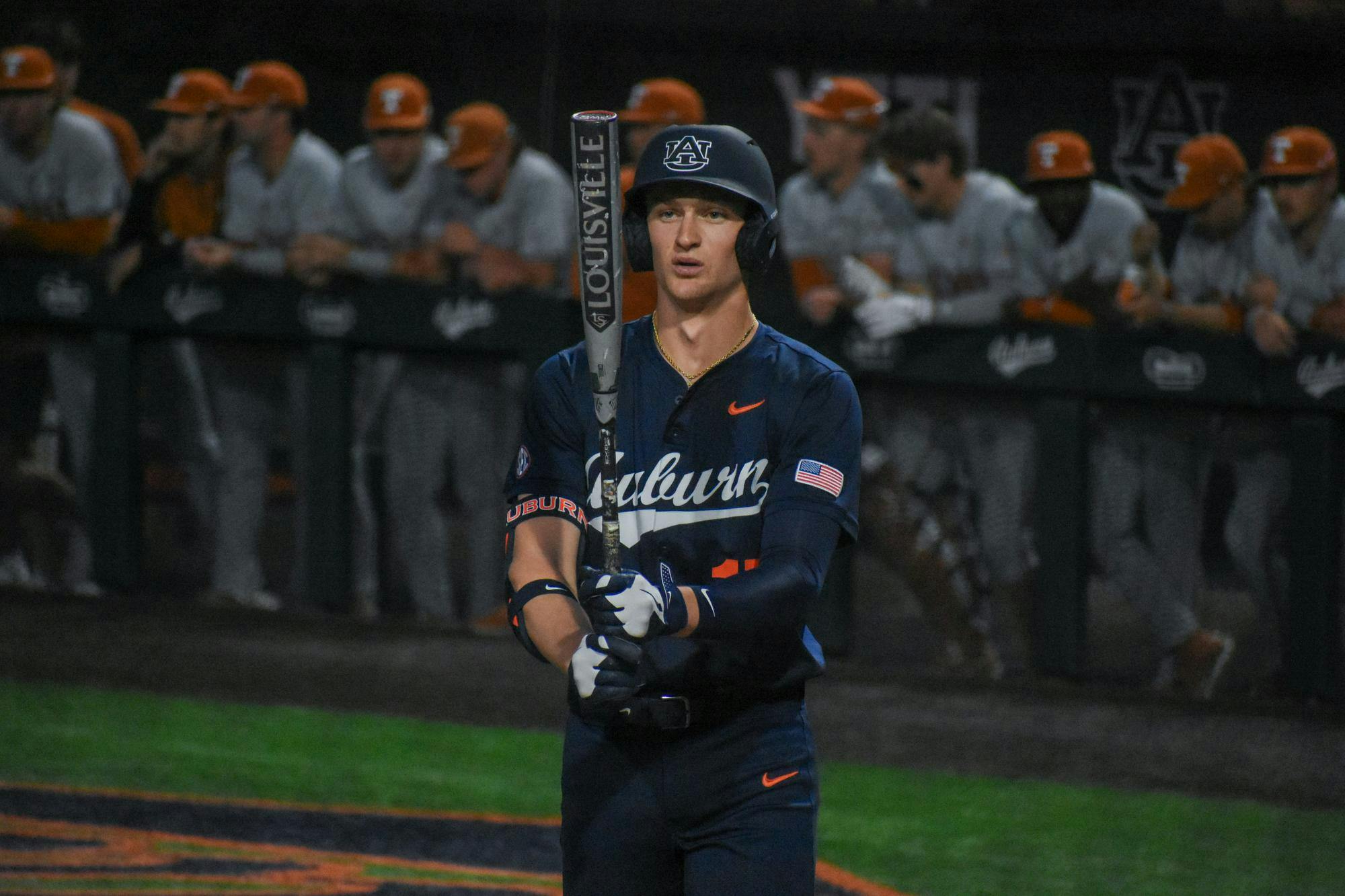 A player in a dark blue baseball uniform holds a bat, focusing intently while teammates watch from the bench in the background.