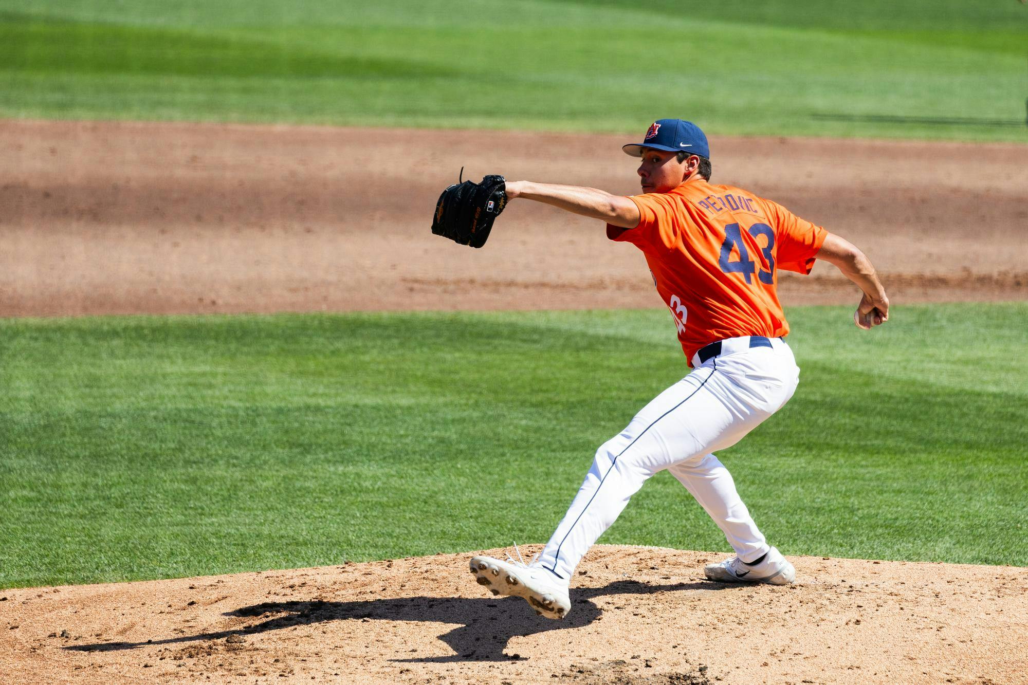 A baseball pitcher in an orange jersey and white pants throws the ball from the mound on a green field.