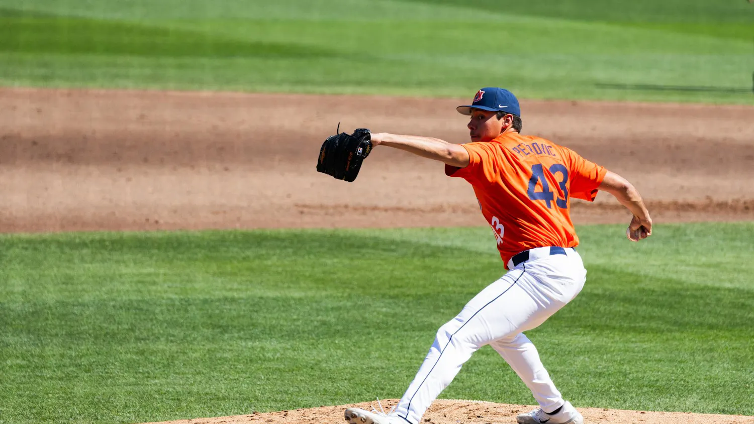 A baseball pitcher in an orange jersey and white pants throws the ball from the mound on a green field.