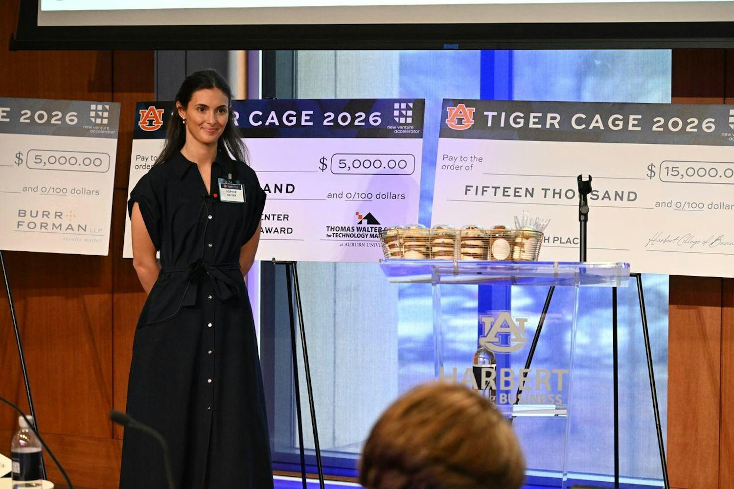A woman in a dark dress stands beside large novelty checks displaying monetary amounts, with desserts arranged on a glass table in front of her.