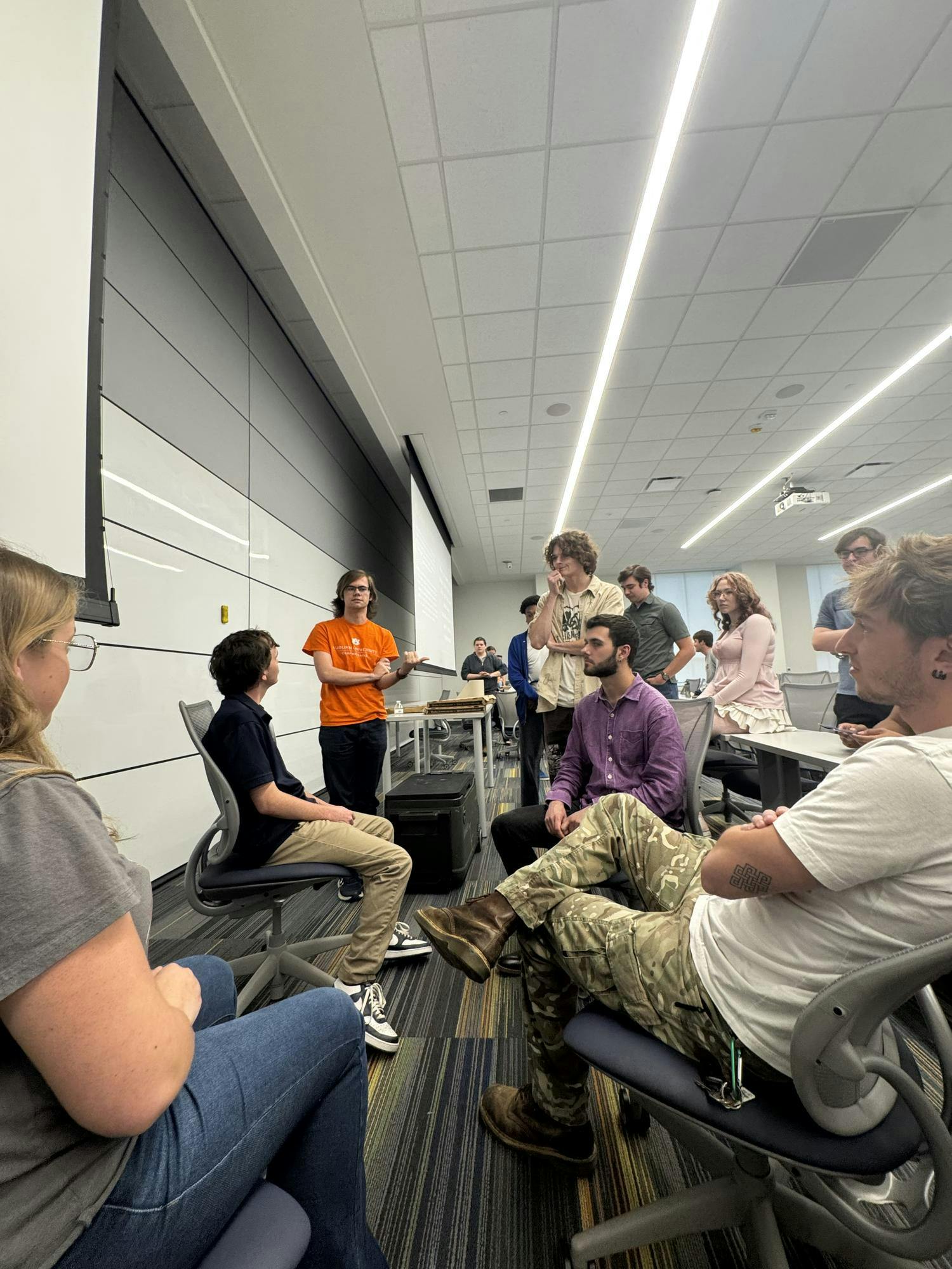 A group of people is engaged in discussion, with one person standing and gesturing while others sit in various positions around them.